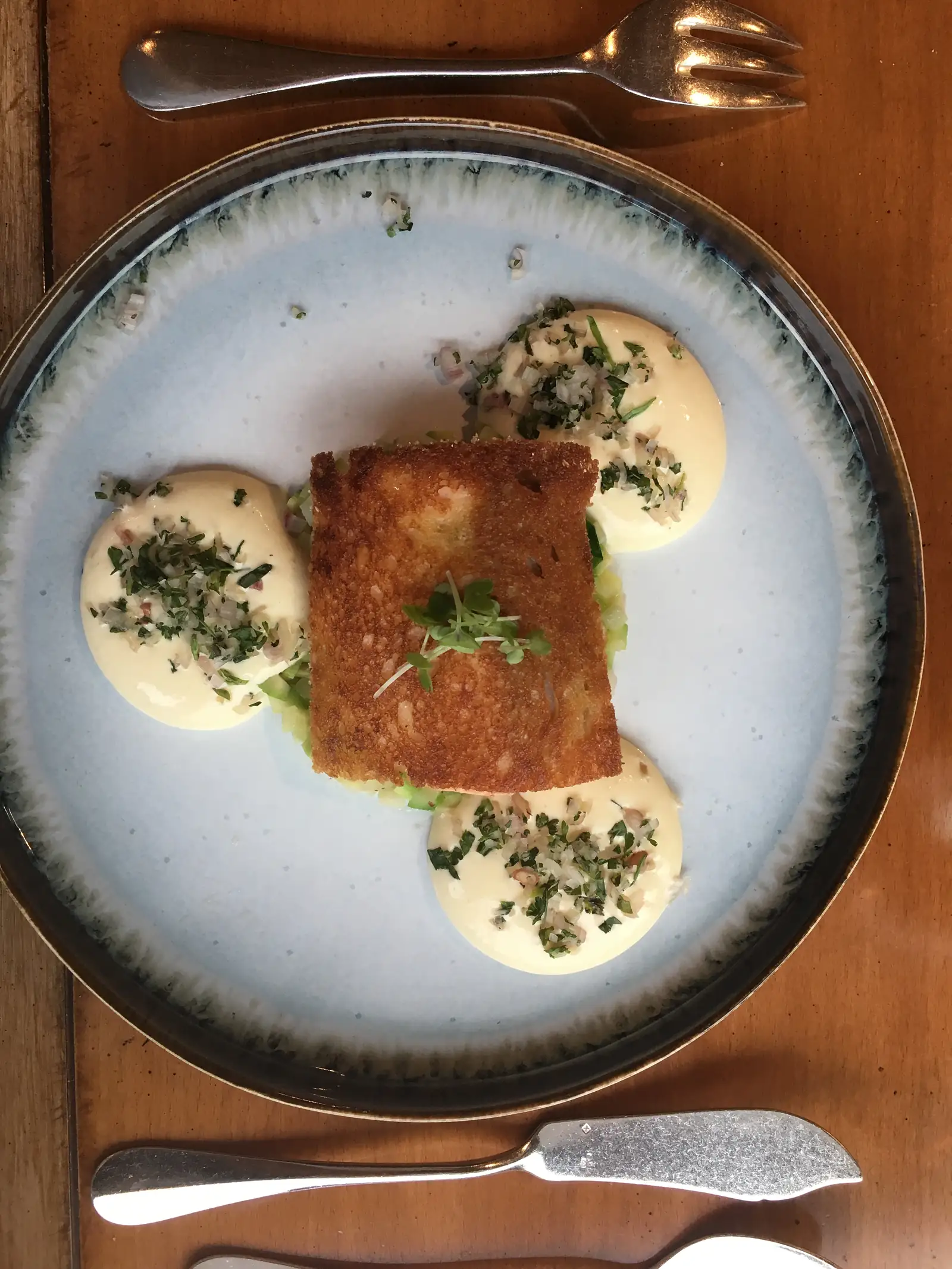 A golden fried fish parcel garnished with cress, plated with three swirls of herb-flecked sauce on a speckled blue-grey plate.