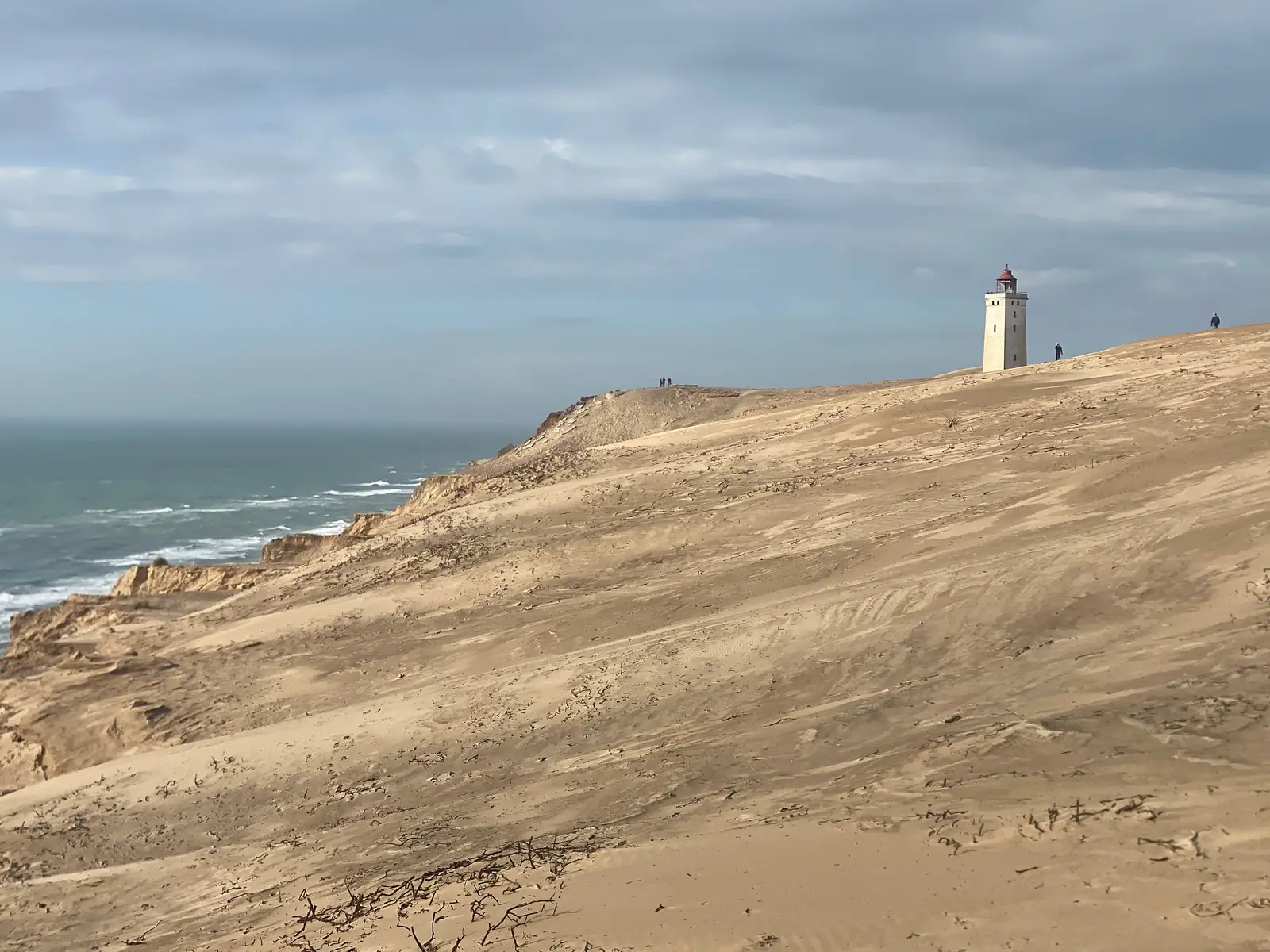 A lighthouse seen along a huge migrating sand dune above eroded cliffs, with tiny figures walking on the ridge.