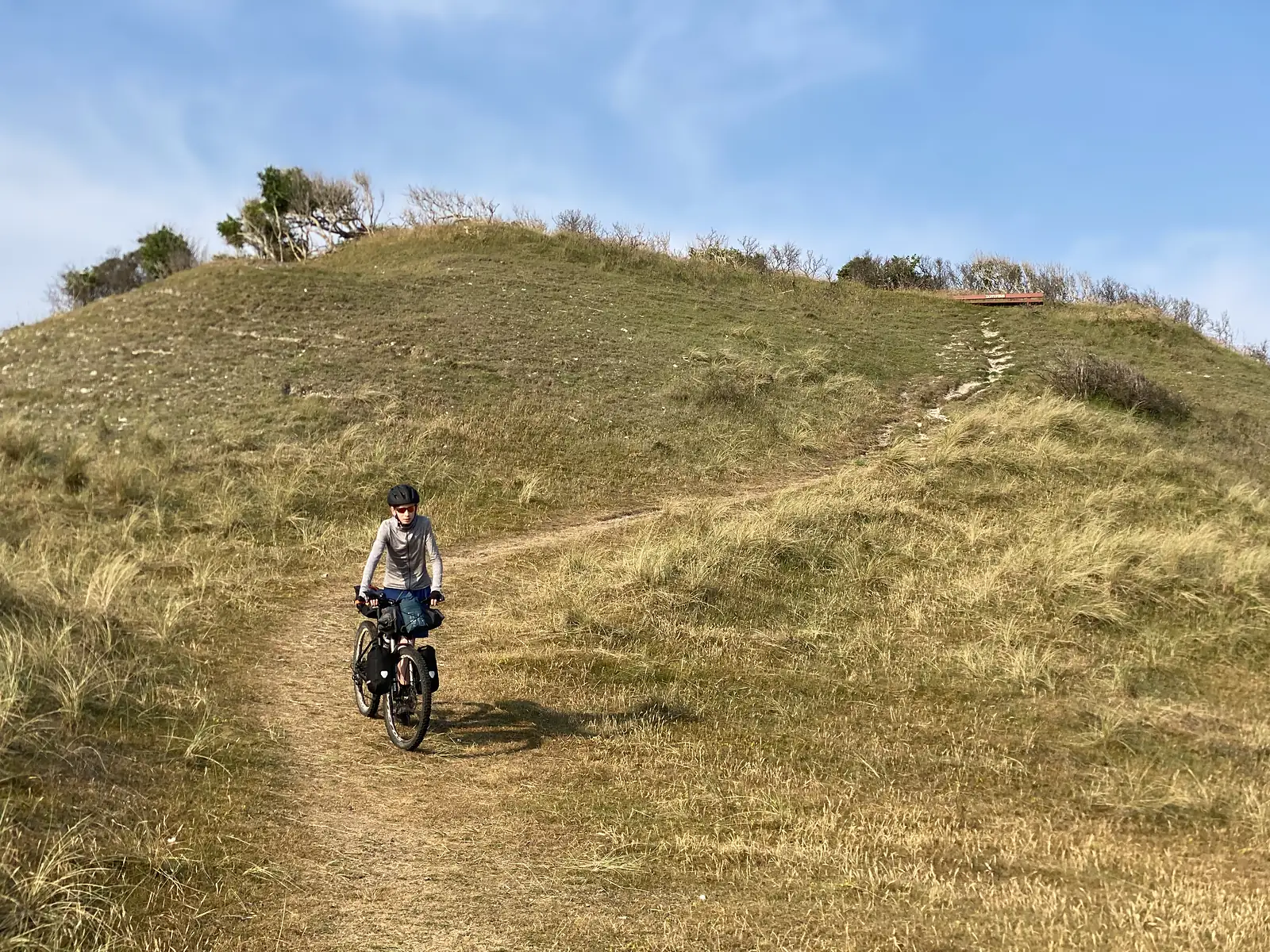 A young cyclist riding a loaded bike down a sandy path from a grassy dune hill, with a wooden viewing platform on the ridge above.