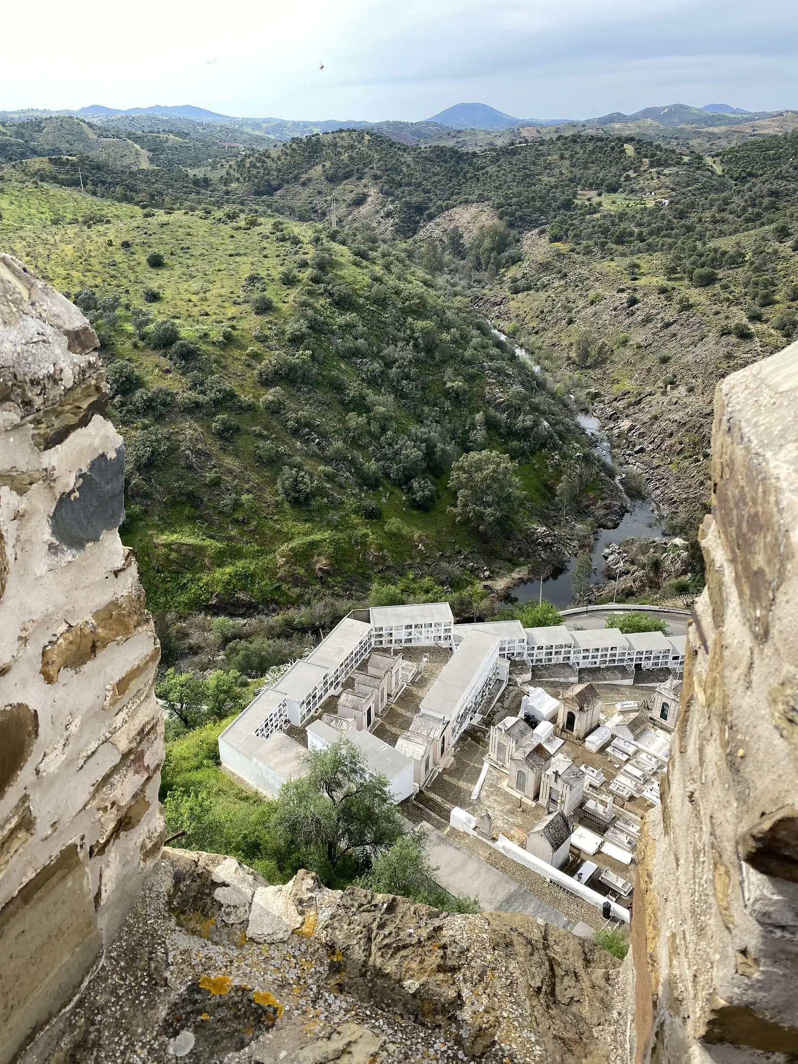 A whitewashed cemetery of tombs and wall niches seen far below through a gap in a castle's stone battlements, with a river winding through a rocky gorge beyond.