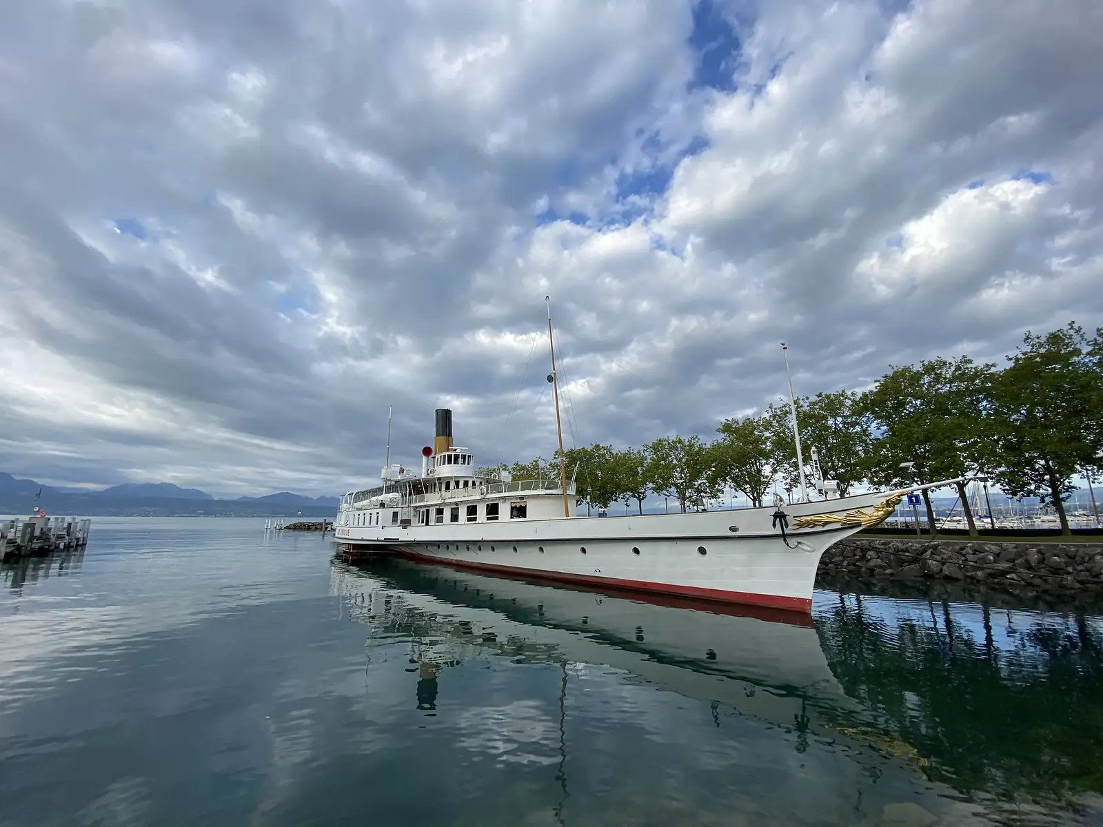 A vintage paddle steamer moored at a lakeside quay, its varnished funnel and gilded bow ornament reflected in the still water beneath a cloudy sky.