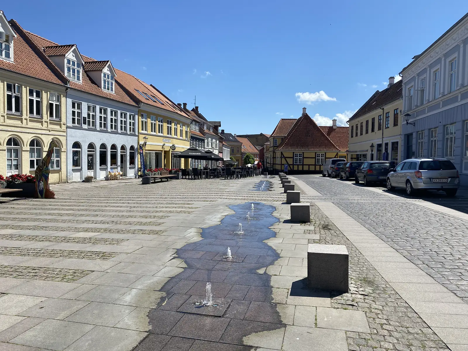 A wide paved town square with a line of small ground fountains, gabled and half-timbered buildings around it and cafe tables under parasols.