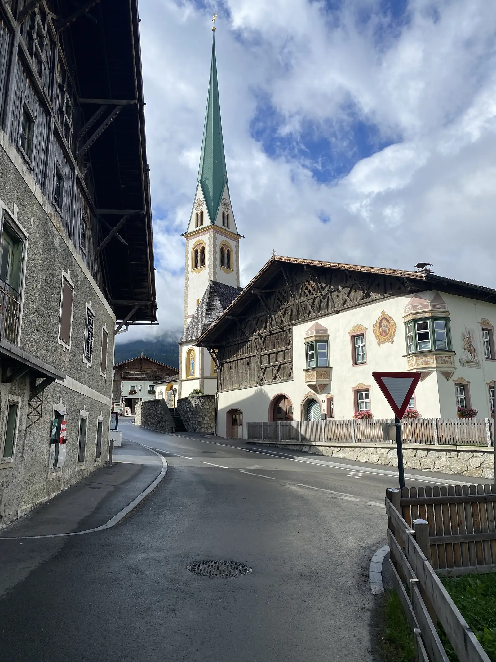 An empty village street runs past a slim green-spired church and a painted alpine house with carved timber gables and a give-way sign.