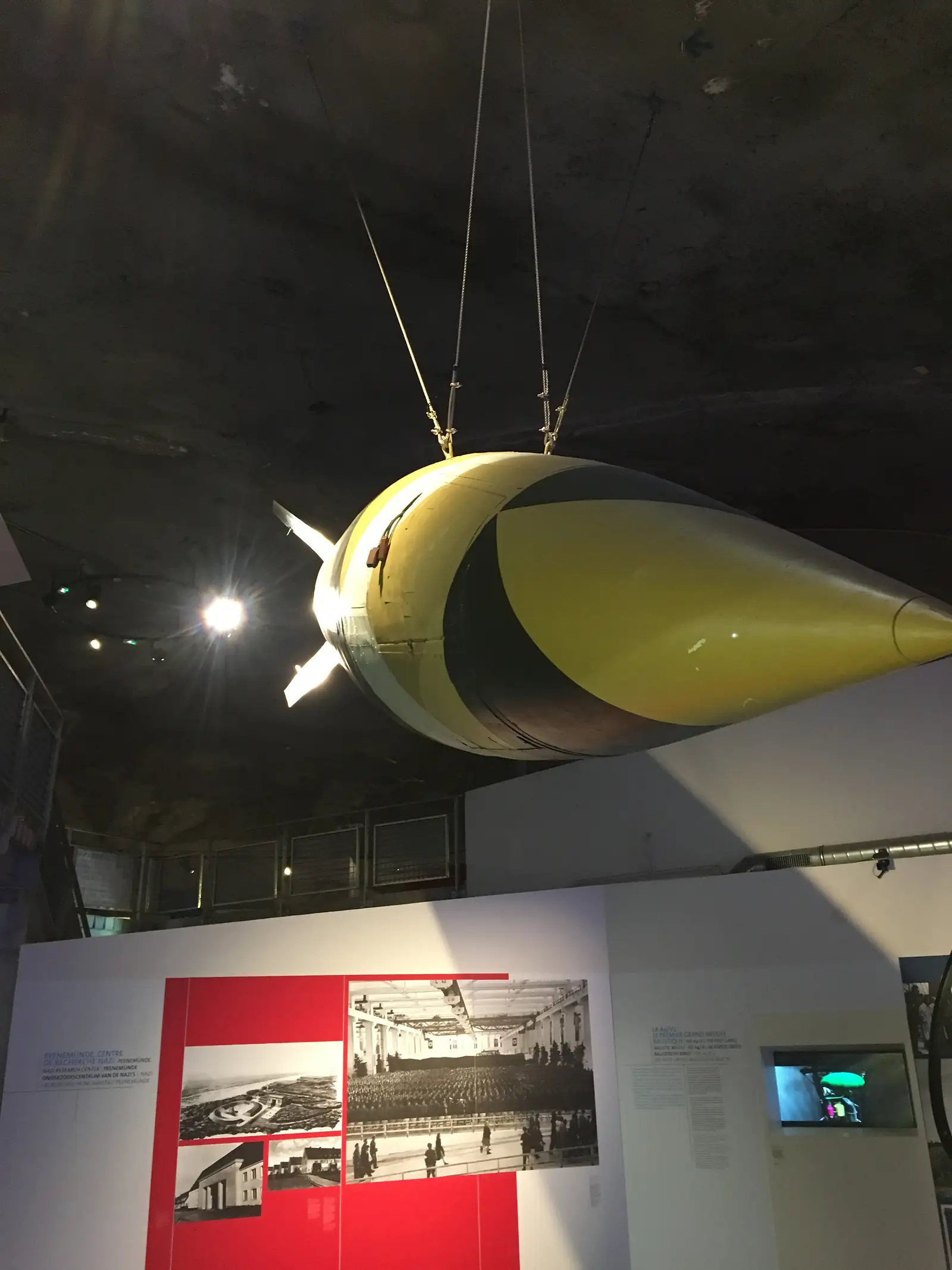 The yellow and black nose of a large bomb-shaped exhibit hangs on cables in a dim hall, with lit displays of archive photographs on the wall behind.