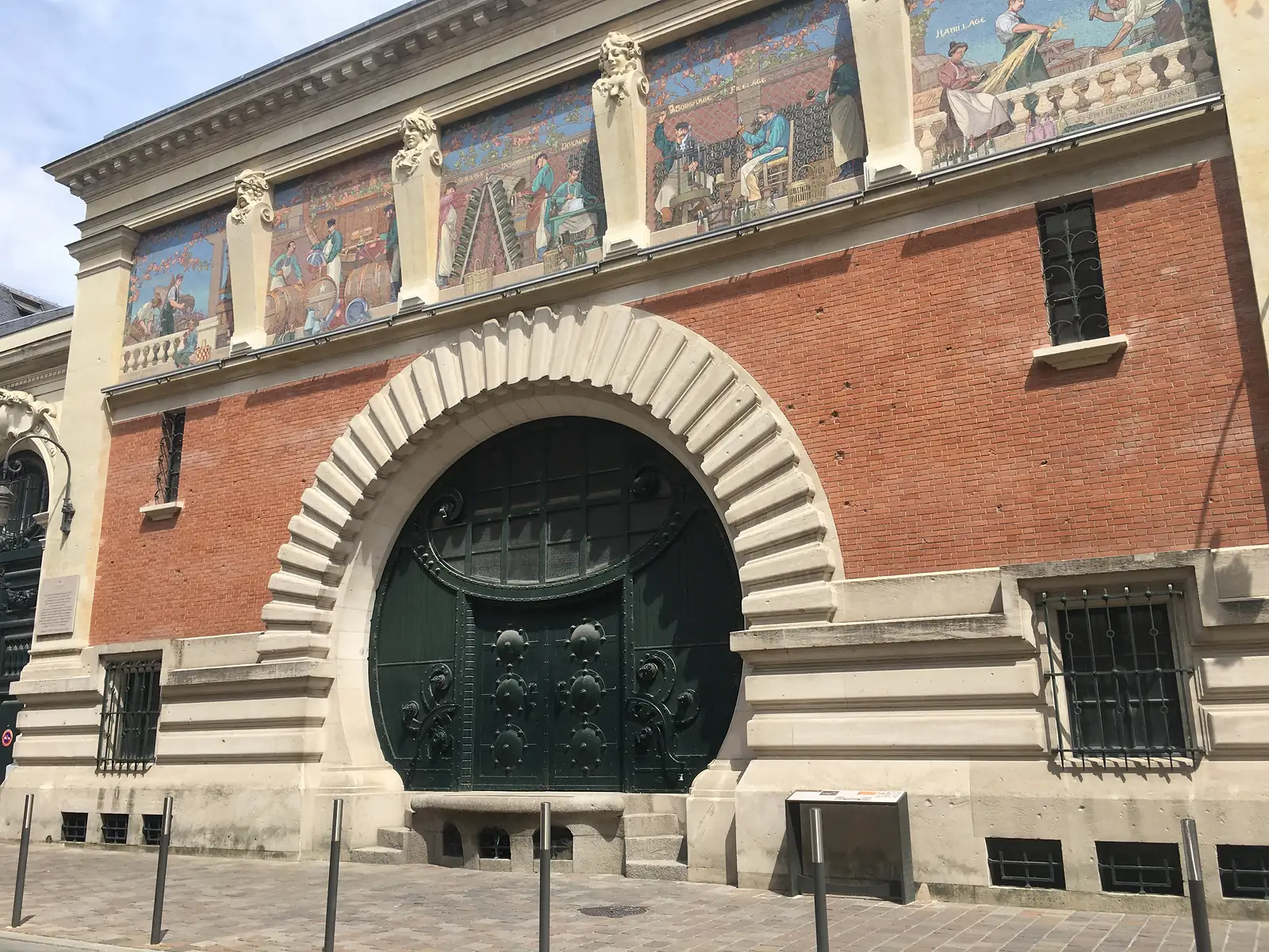 A round-arched iron door set in red brick sits beneath a mosaic frieze depicting Champagne-making scenes labelled HABILLAGE and BOUCHAGE FICELAGE DOSAGE.