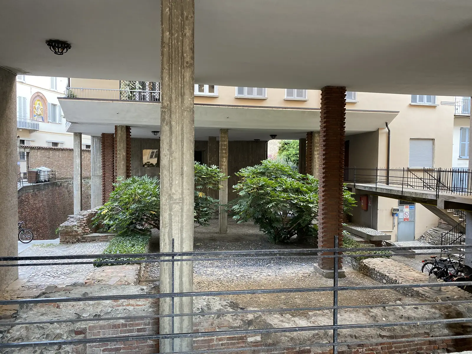Brick columns and exposed medieval foundations frame a quiet courtyard garden shaded by ferny shrubs.