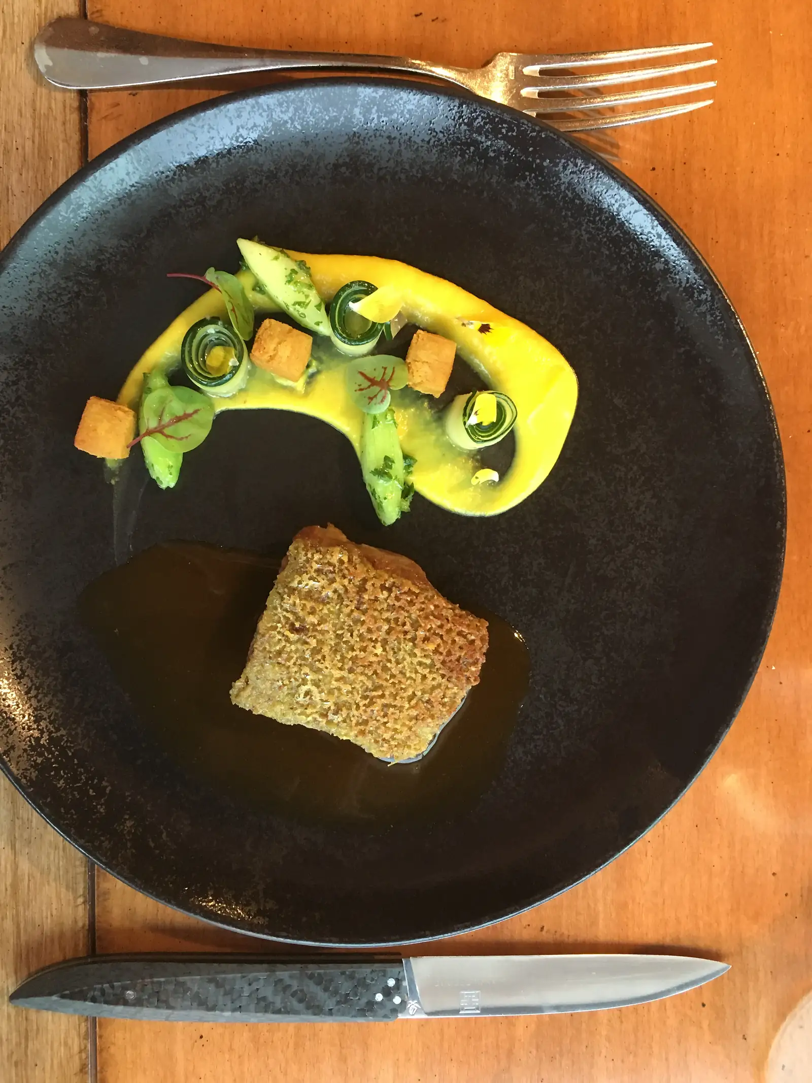 A breadcrumb-crusted portion of meat in dark jus beside a swirl of yellow purée, courgette rolls and croutons, plated on a matte black dish.