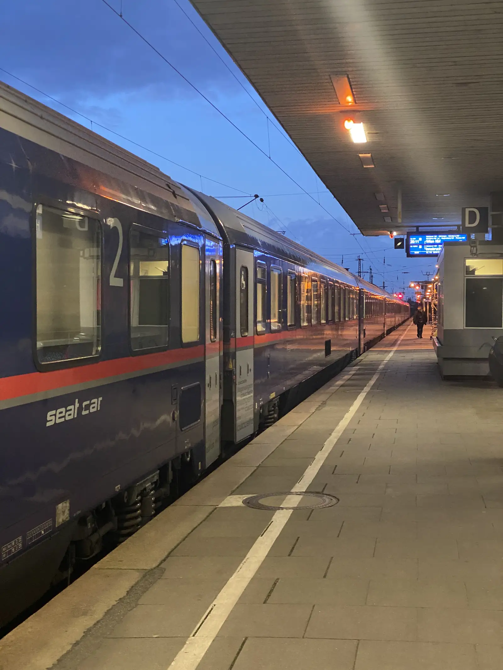 A dark blue sleeper carriage marked 'seat car' stands at a covered platform in the evening light.
