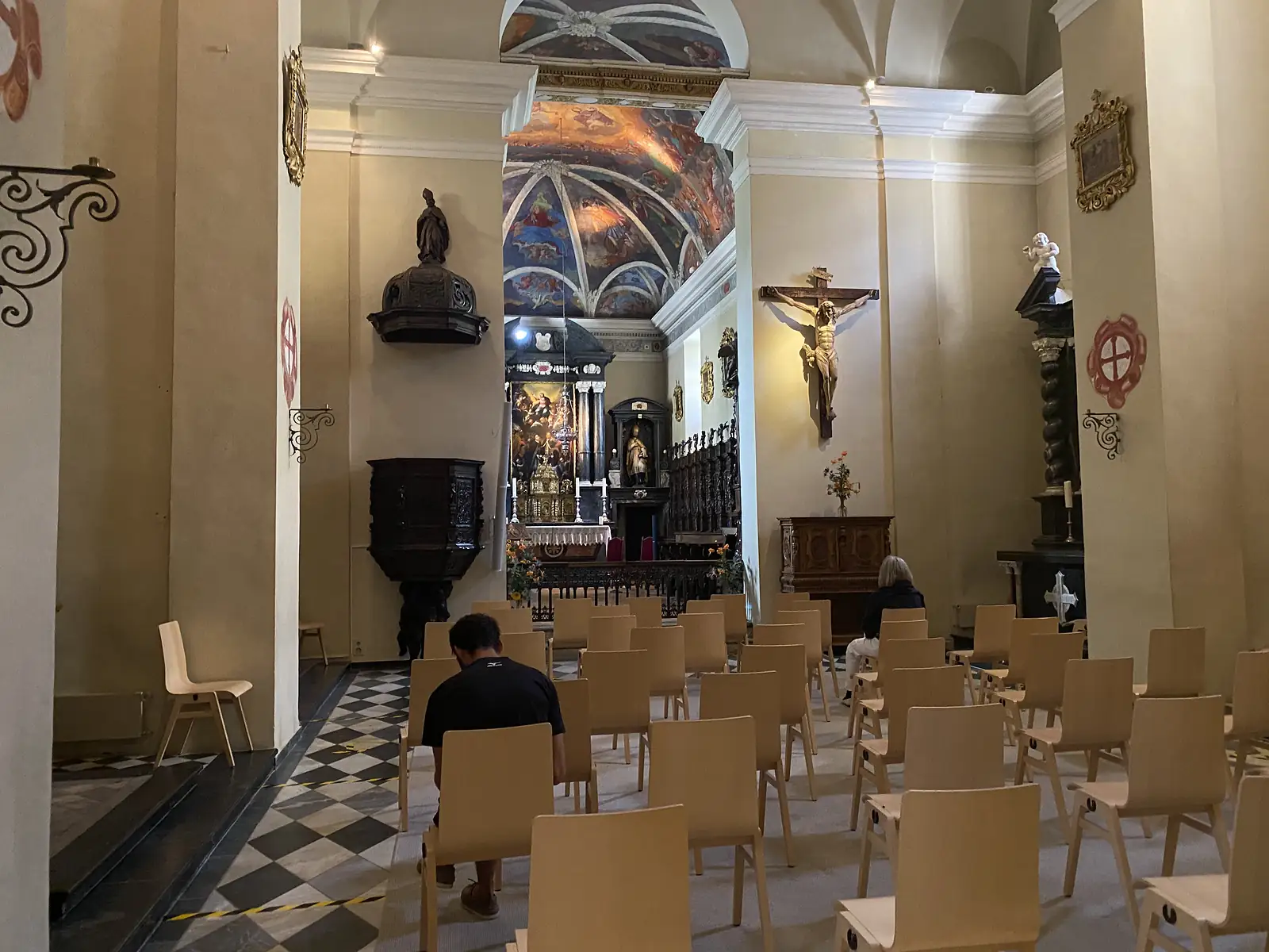 The nave of a baroque church looking towards a gilded altar beneath a frescoed dome, with a few visitors seated among rows of wooden chairs.