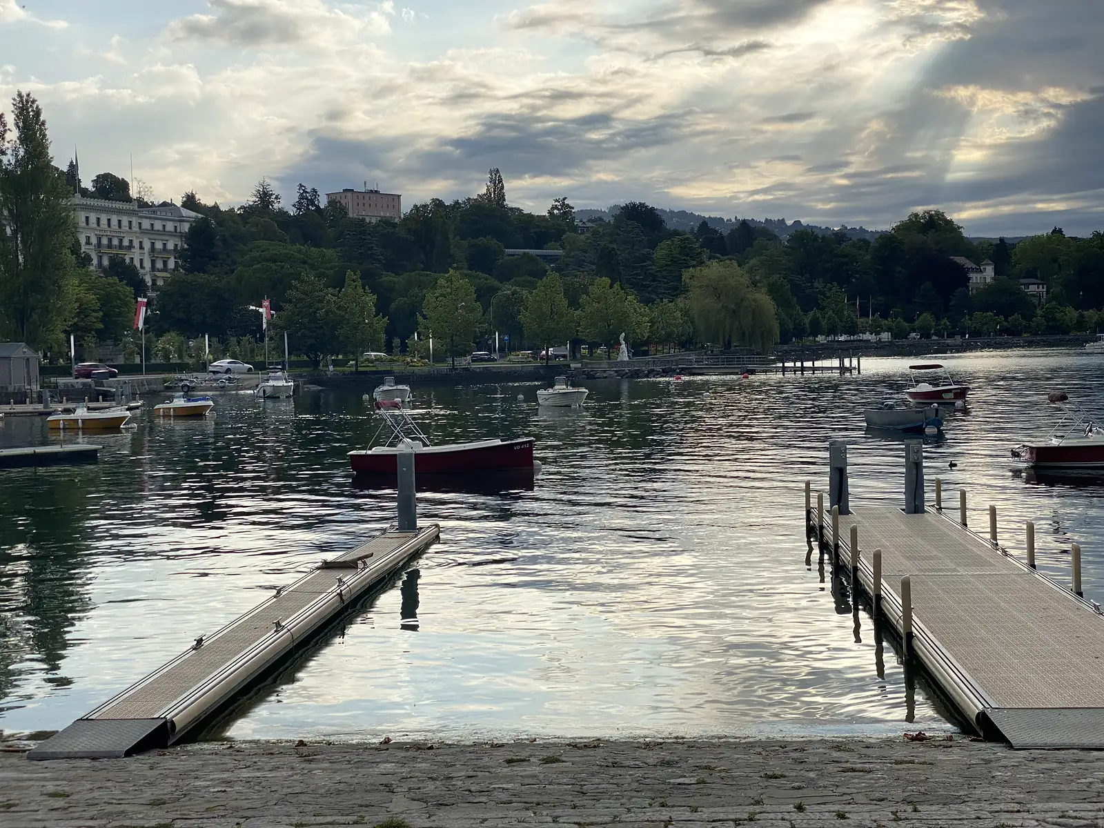 A small marina of moored motorboats at dusk, with grand lakeside buildings and wooded hills rising behind the water.