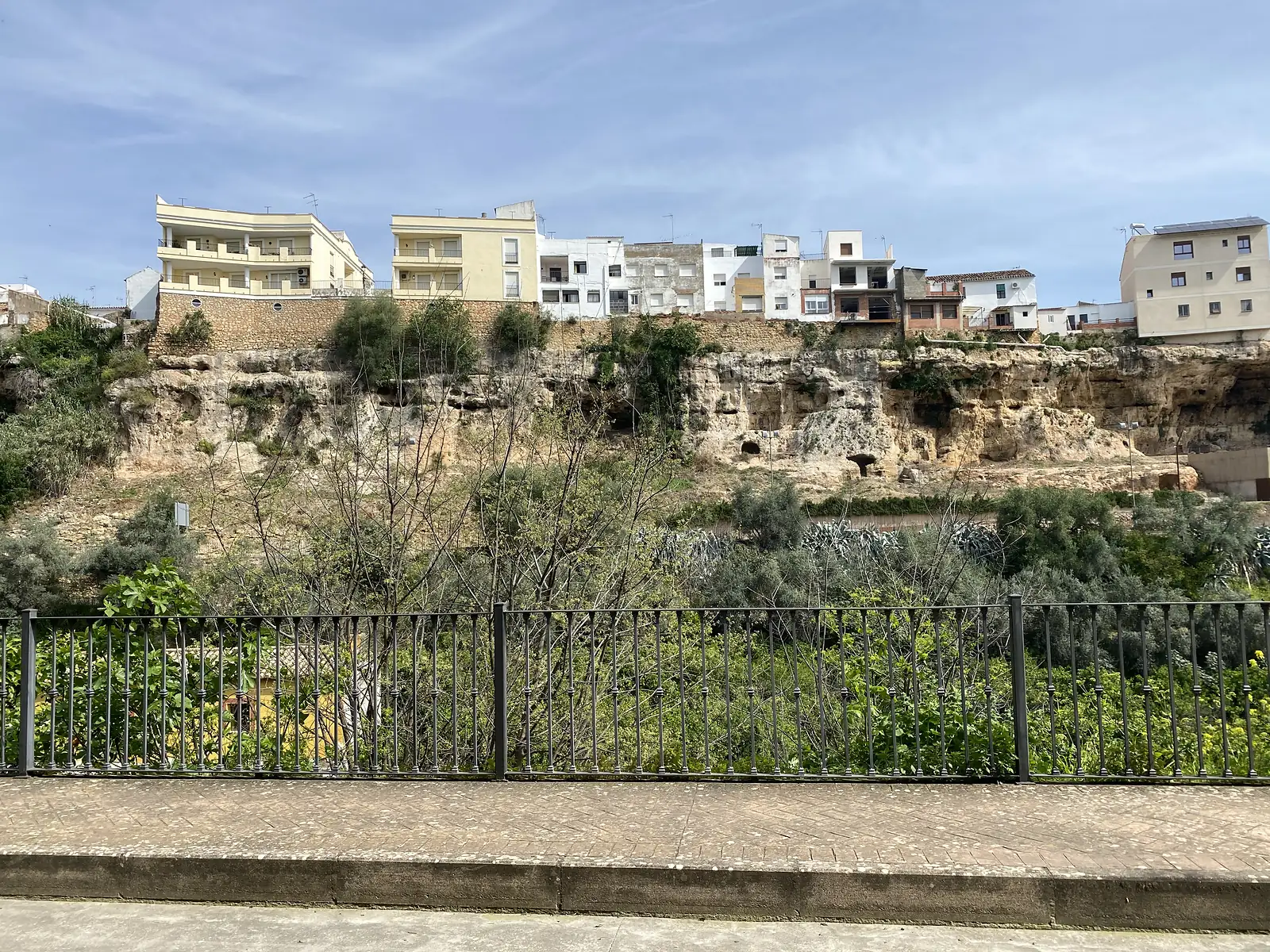 Houses lining the top of a limestone cliff riddled with cave openings, seen across a green ravine from behind roadside railings.