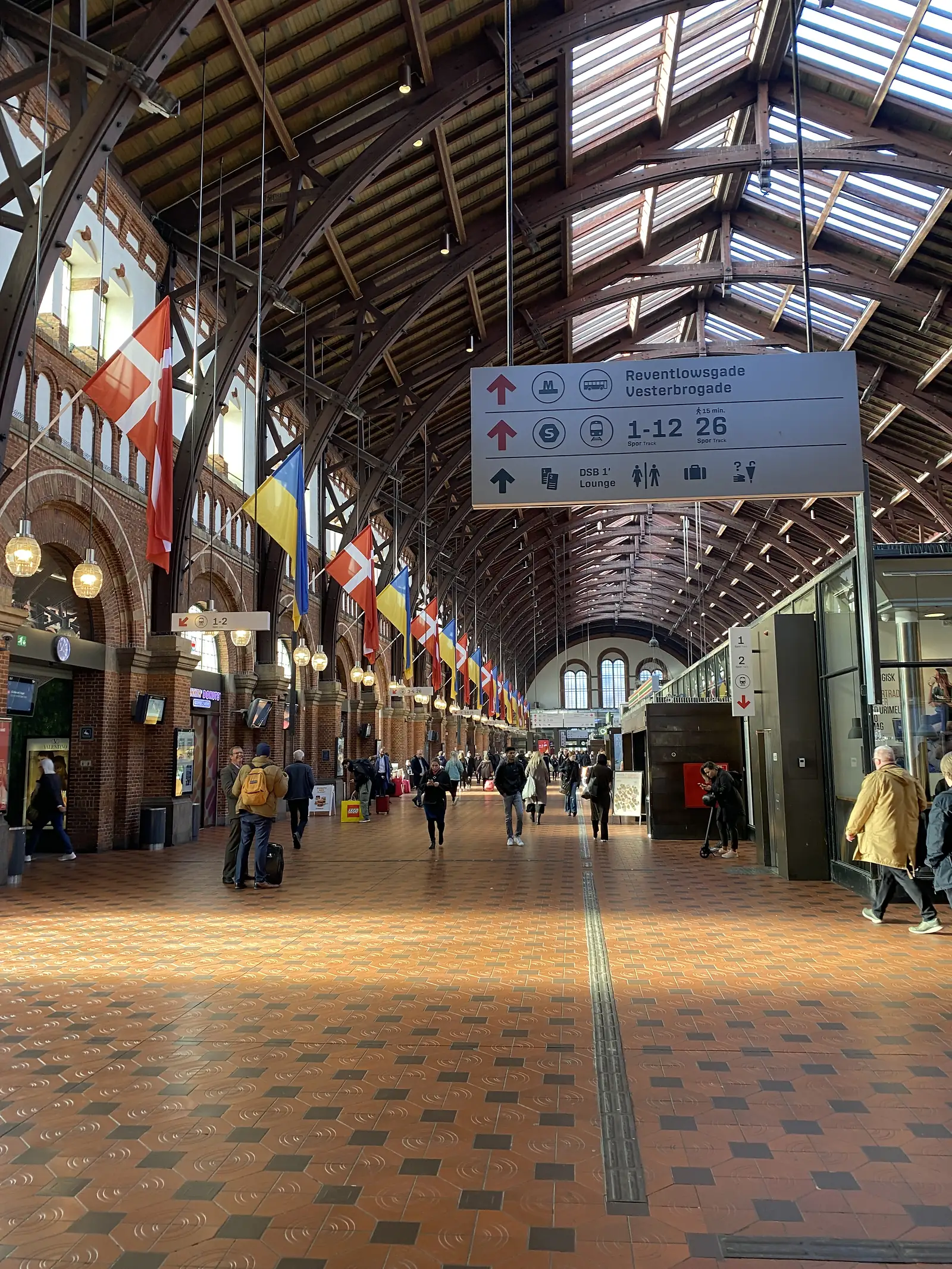 Danish flags hang from the vaulted roof of a grand railway station hall, with a platform sign reading 'Reventlowsgade Vesterbrogade, 1-12, 26'.
