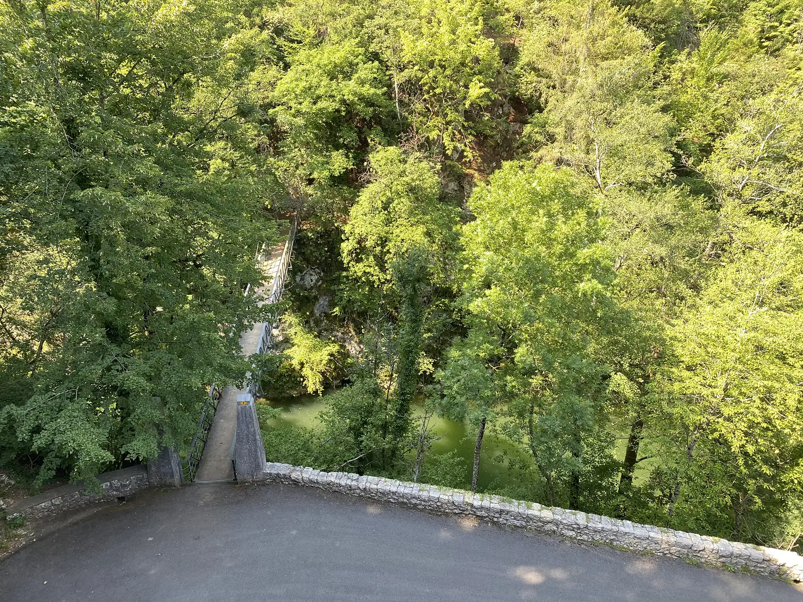 A stone parapet bridge viewed from above, with a narrow footbridge crossing a tree-shaded gorge and a green river below.
