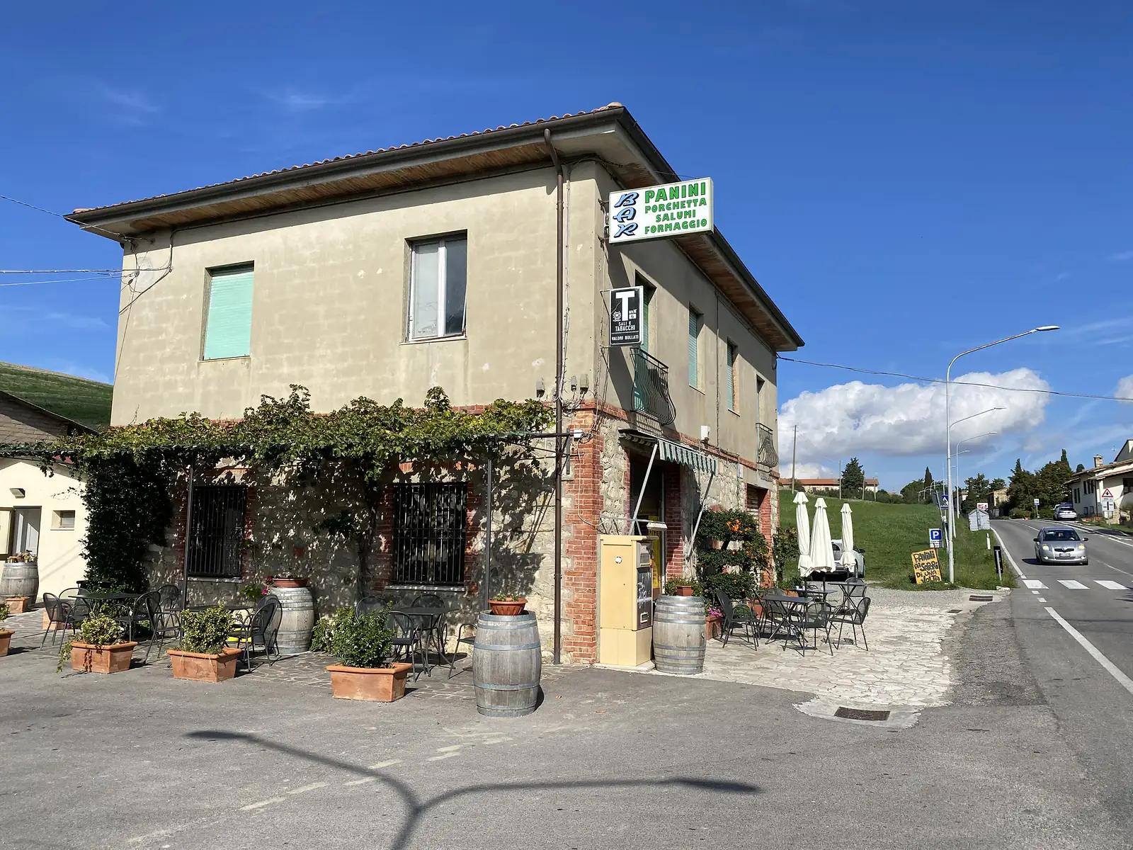 A stone and stucco bar on a corner in Tuscany, its facade covered in climbing vines, with a green-lettered 'PANINI PORCHETTA SALUMI FORMAGGIO' sign, wine barrels and outdoor tables on the pavement, and folded umbrellas beside the entrance.