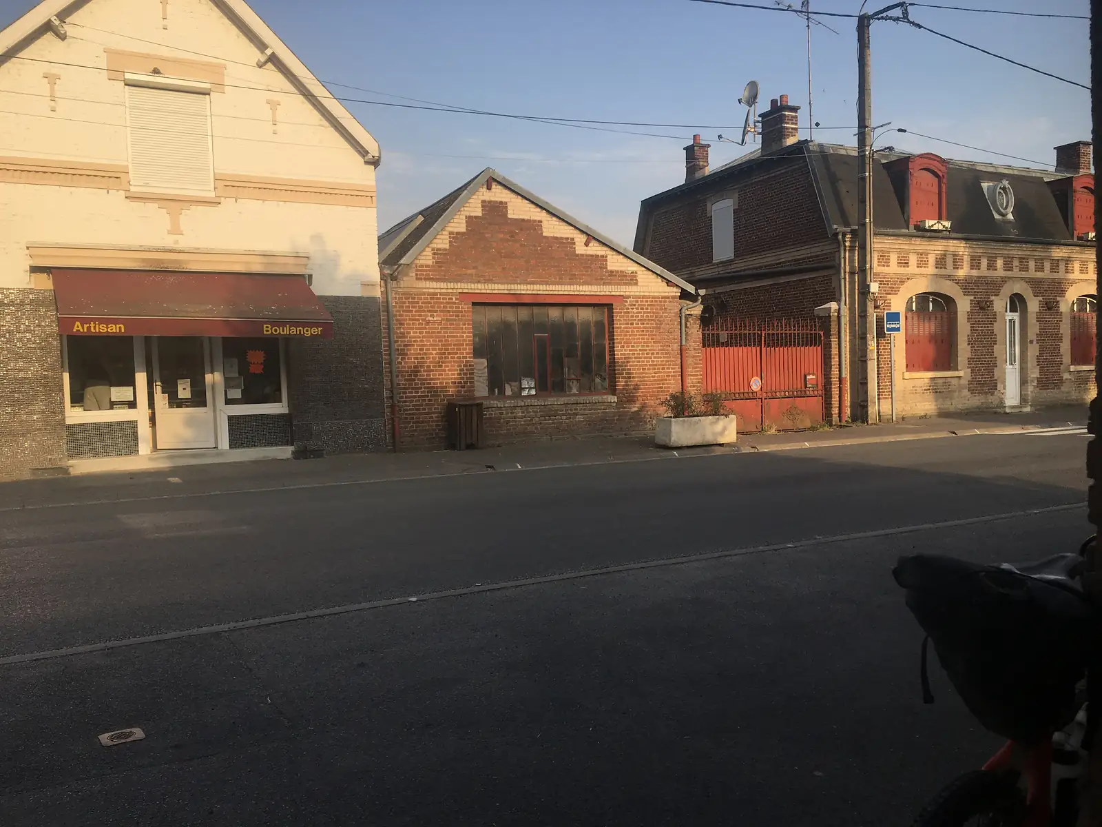 A village bakery with a red awning reading 'Artisan Boulanger' sits on an empty street corner, a loaded touring bike visible at the edge of the frame.
