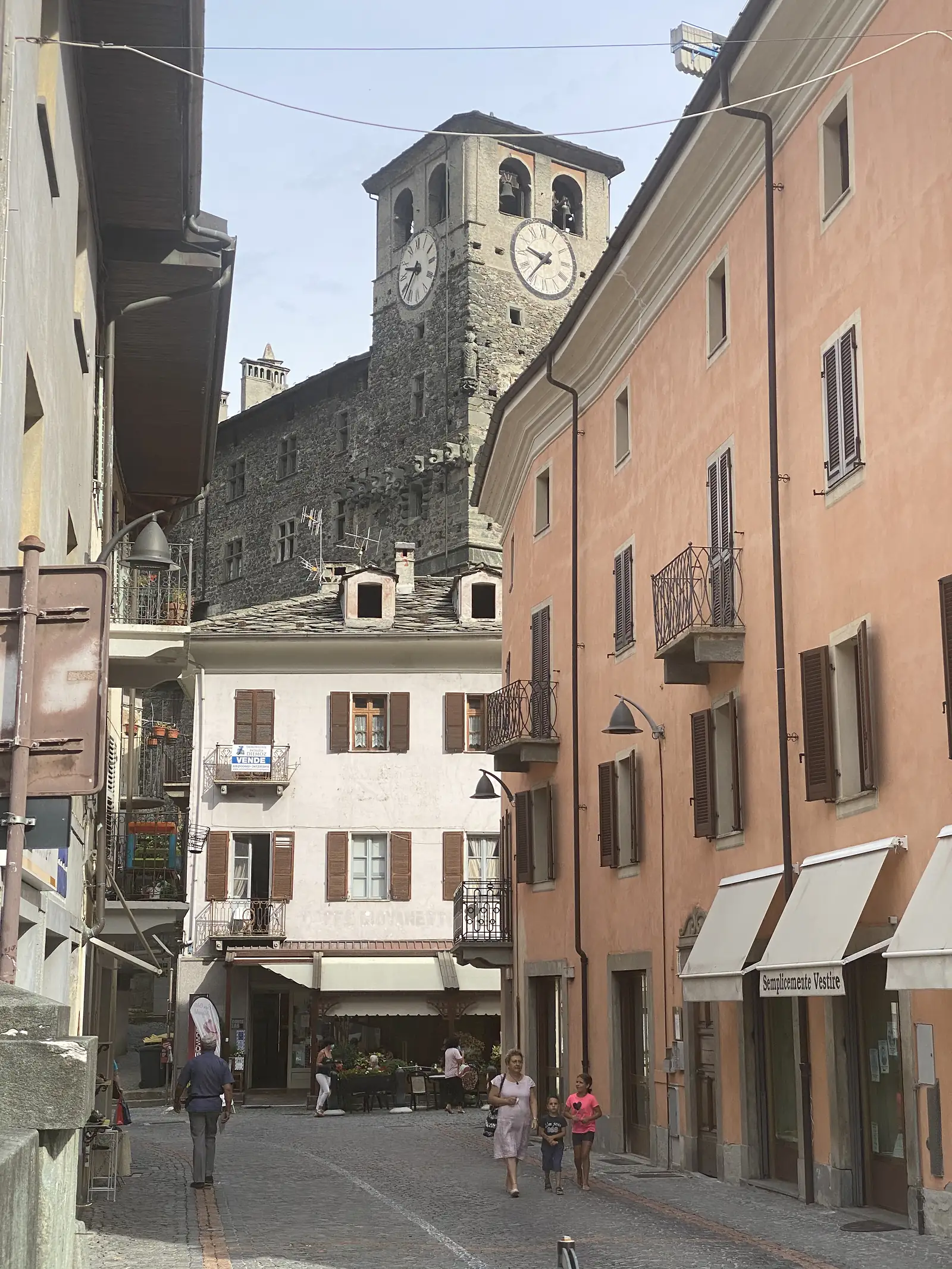 A narrow street leading towards a stone clock tower with two dial faces, flanked by a white-walled house and a coral-pink apartment building, with pedestrians including two children walking past.