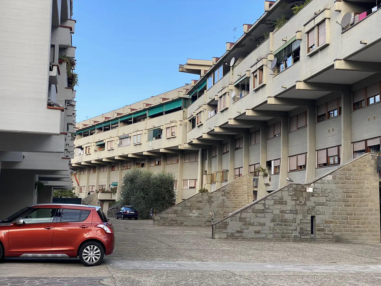 A tiered concrete apartment block with stepped terraces and external stairways overlooking a courtyard where cars are parked.