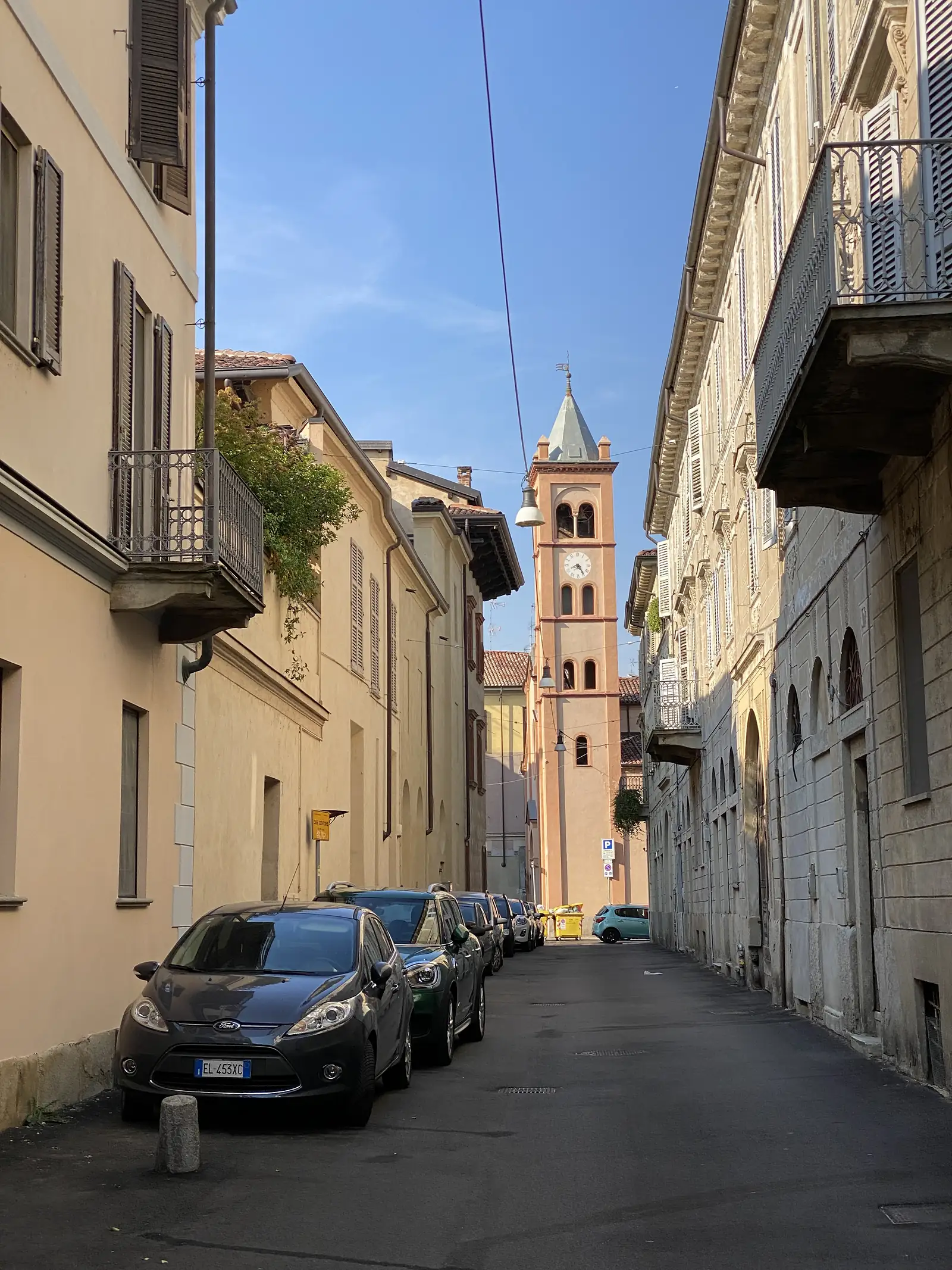 A narrow street in Vercelli lined with shuttered buildings, cars parked along the kerb and a brick clock tower standing at the end of the road.