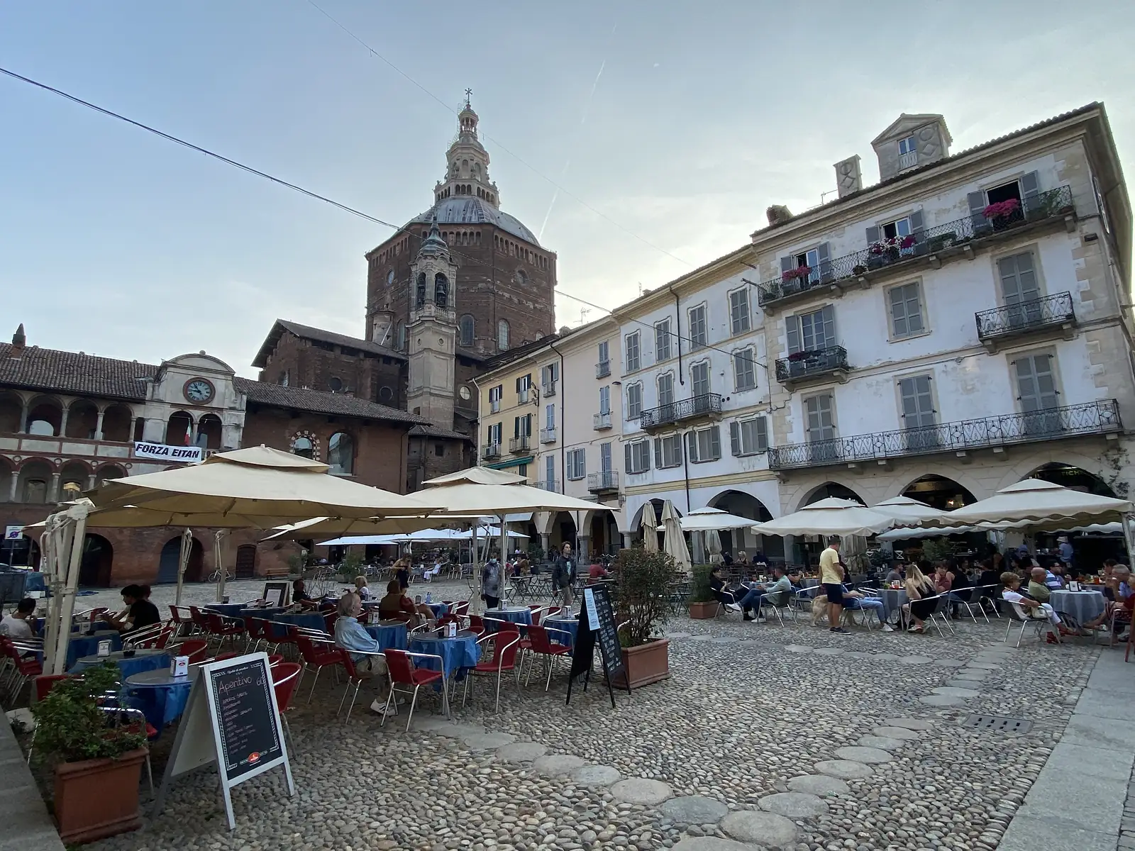 Diners fill café tables under cream parasols in a cobbled square, a banner reading 'FORZA EITAN' strung above the crowd and the brick dome of Pavia's cathedral rising behind.