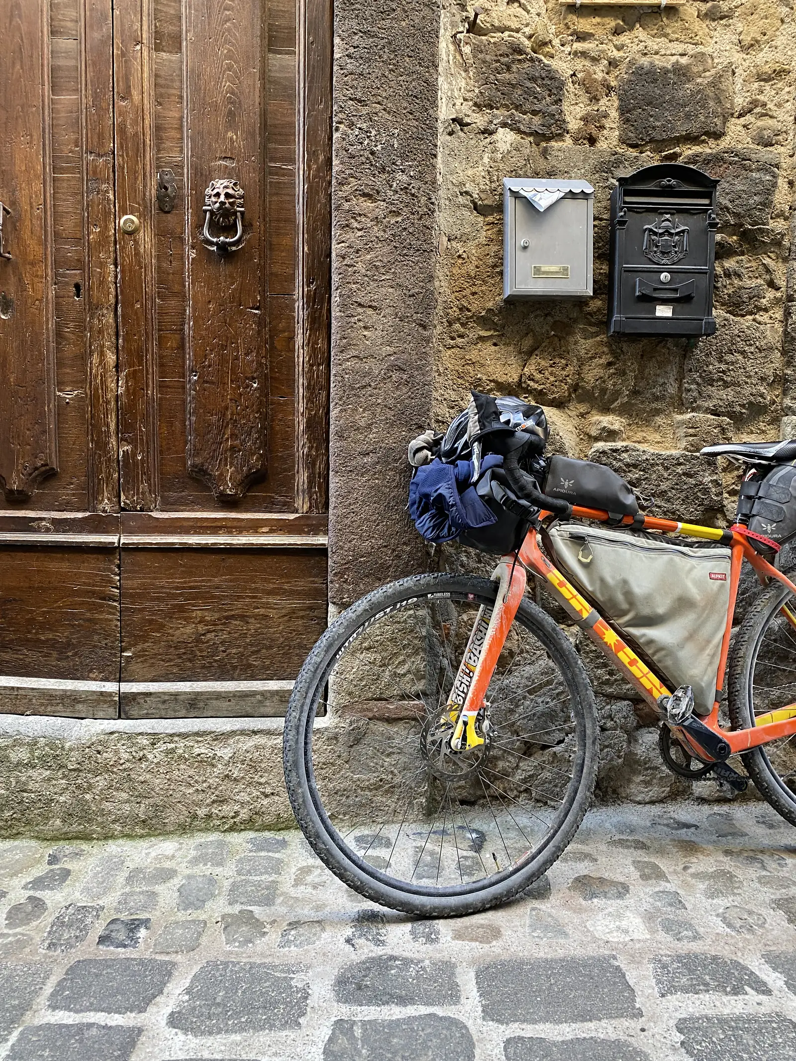 A loaded orange bikepacking bike leans against a stone wall beside letterboxes and an old wooden door with a lion's-head knocker.