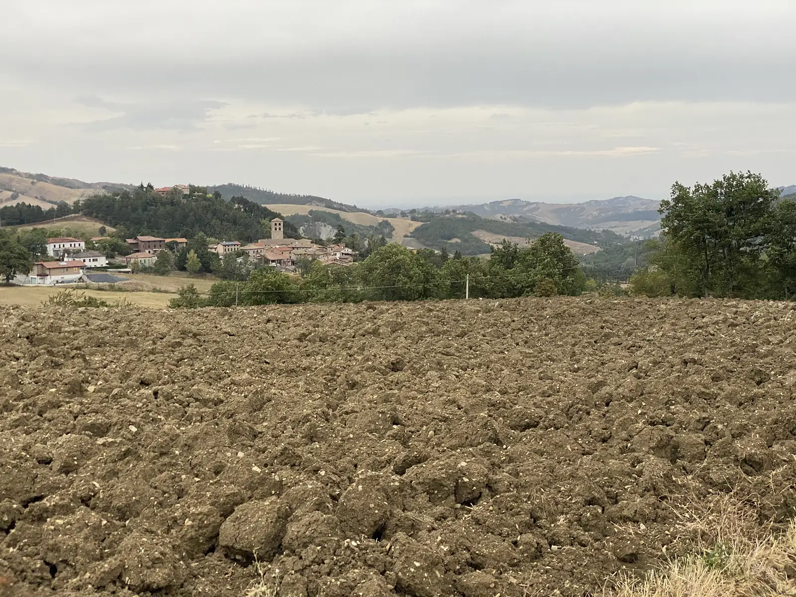 A freshly ploughed field falls away towards a small hilltop village with a bell tower, seen beneath a bank of grey cloud.