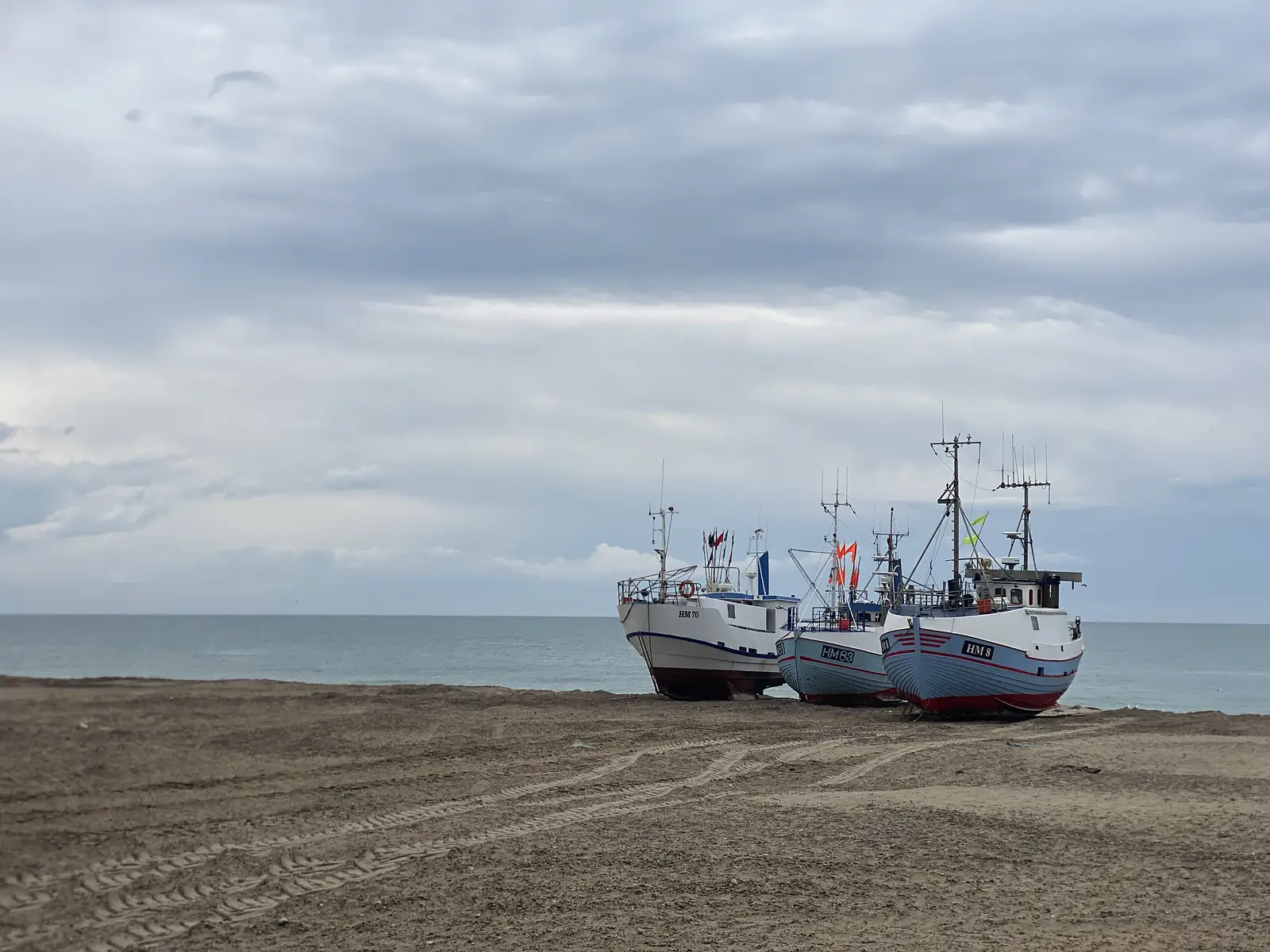 Three beach-launched fishing boats lettered "HM70", "HM63" and "HM8" drawn up on the sand at the water's edge under a heavy grey sky.