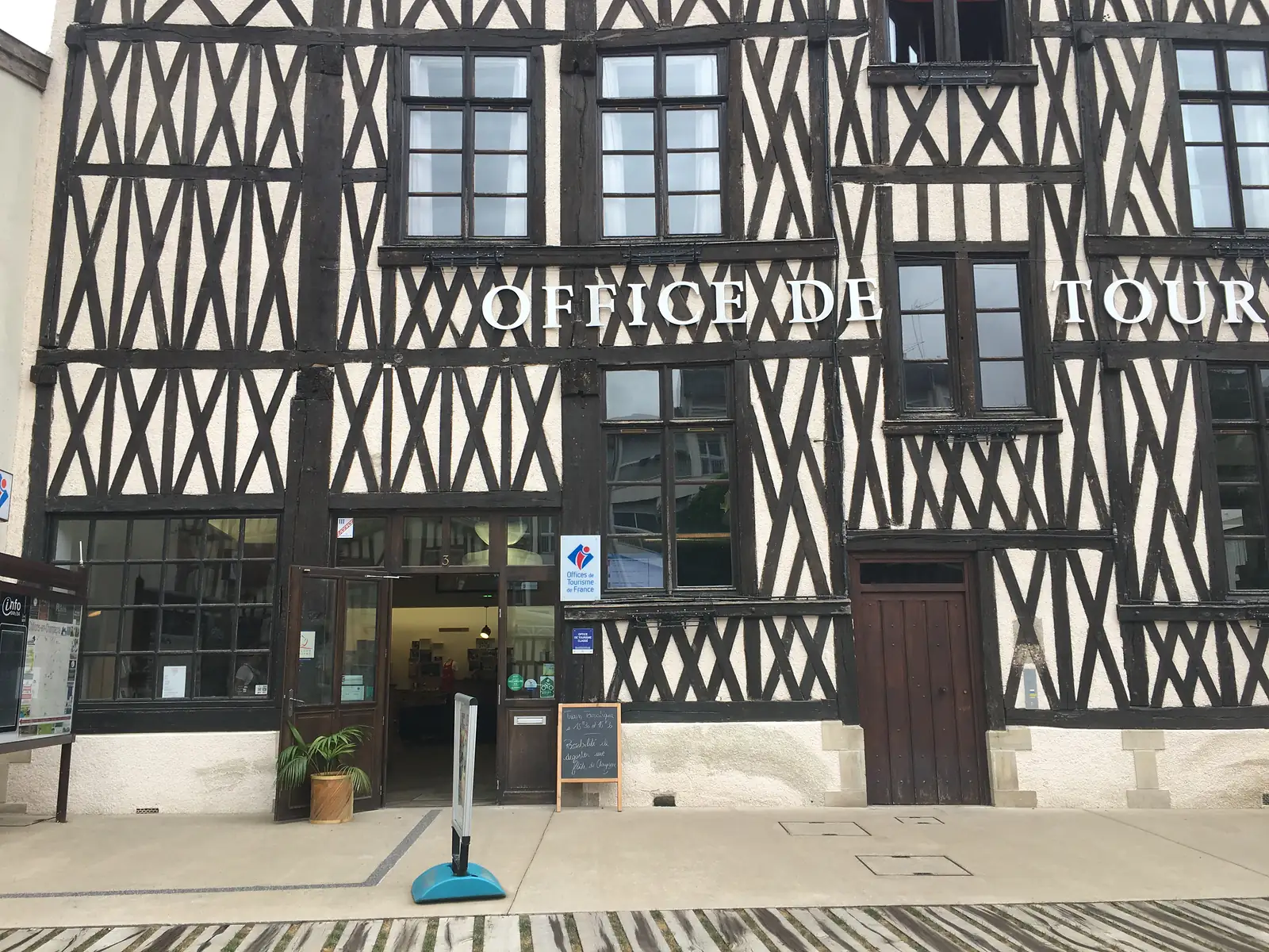 Entrance to a half-timbered tourist office building, its facade lettered 'OFFICE DE TOUR[ISME]' above open double doors and a chalkboard sign on the pavement.