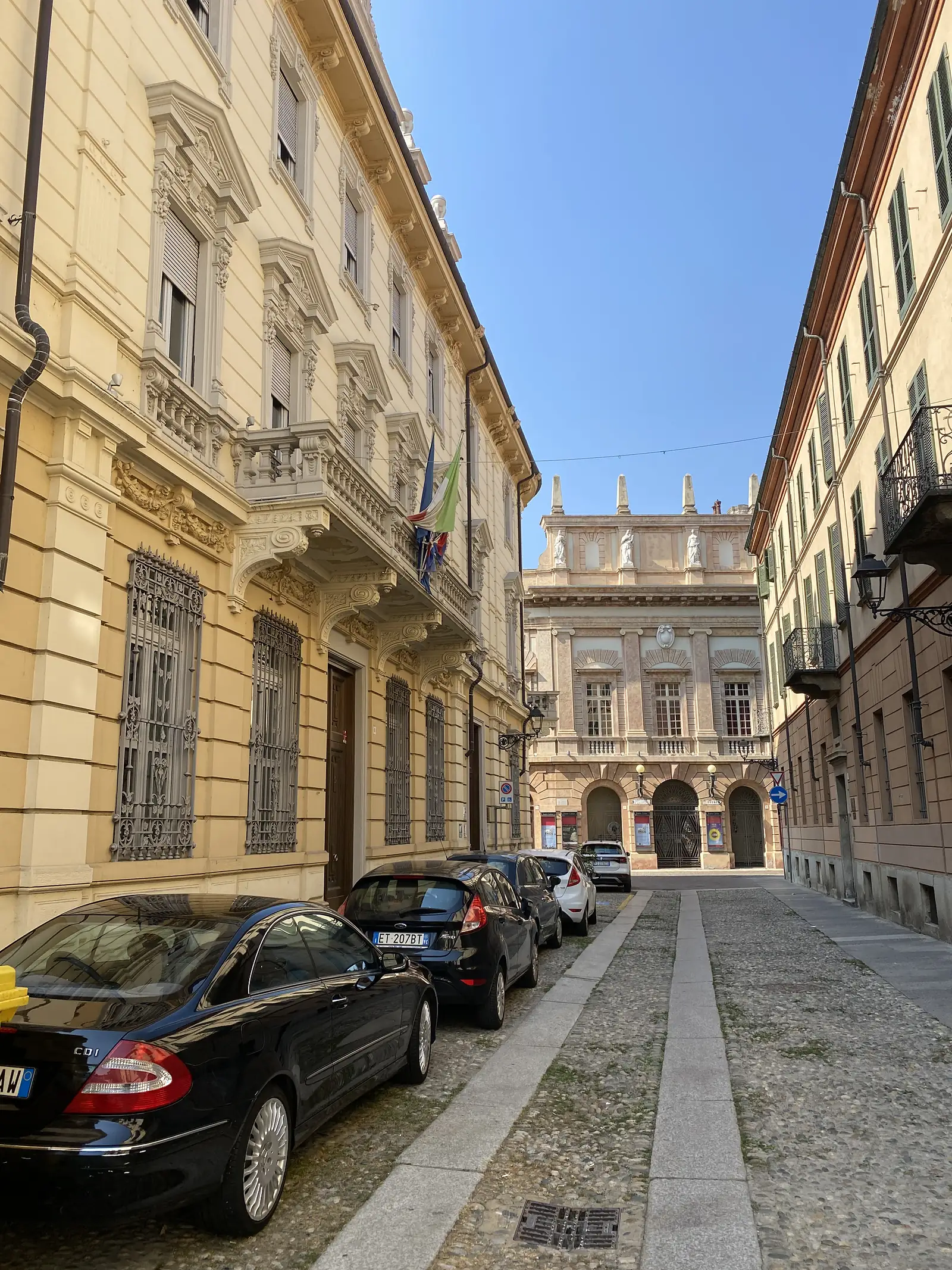 An ornate cream-coloured townhouse facade with wrought-iron balconies and a small Italian flag, cars parked along the cobbled street below.