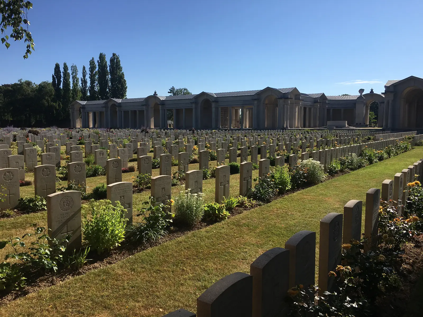 A view along the curved colonnade of the Arras Memorial, its arches framing a stone memorial altar with a wreath, headstones lined up in the foreground.