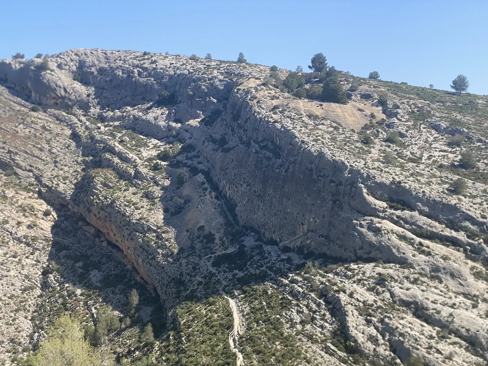 A great grey limestone cliff with a deep overhanging hollow, a stepped path running down beside it on the bare mountainside.
