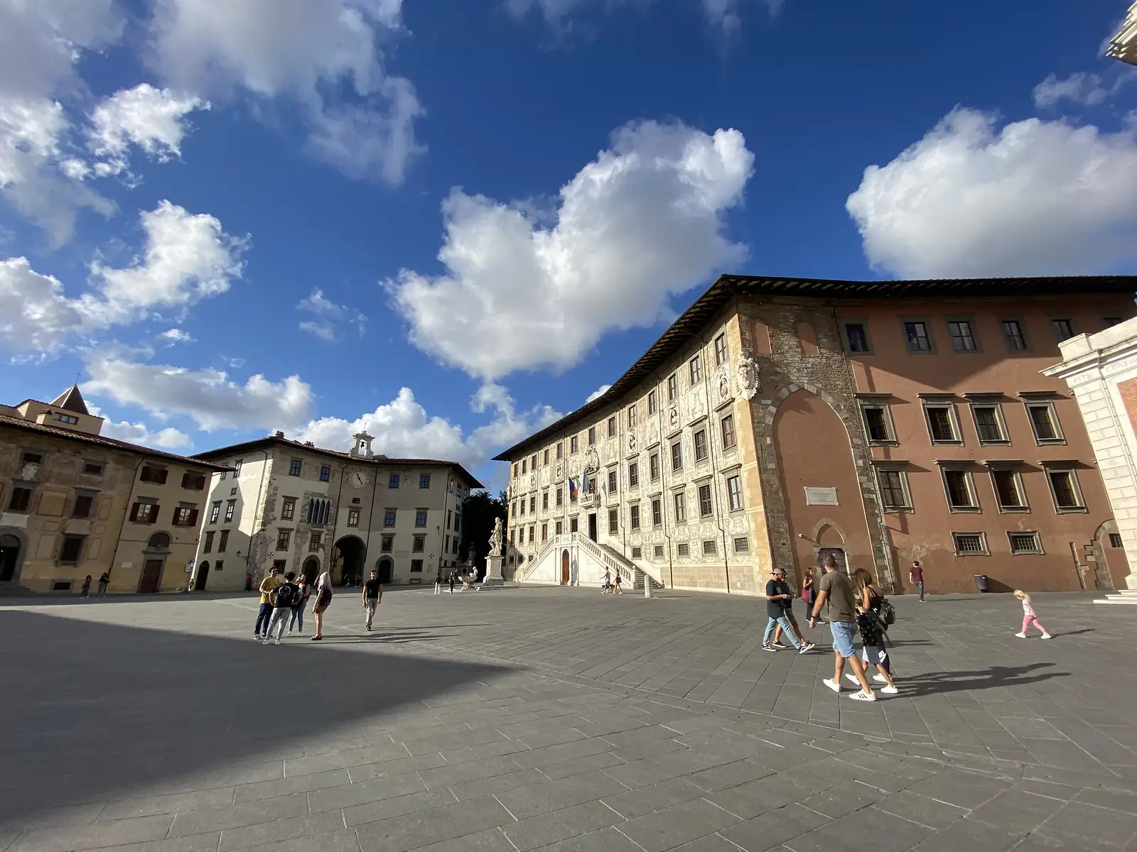 People walking across a broad piazza past an ornately decorated Renaissance palazzo with a scalloped roofline, a marble statue on a plinth in front of it.
