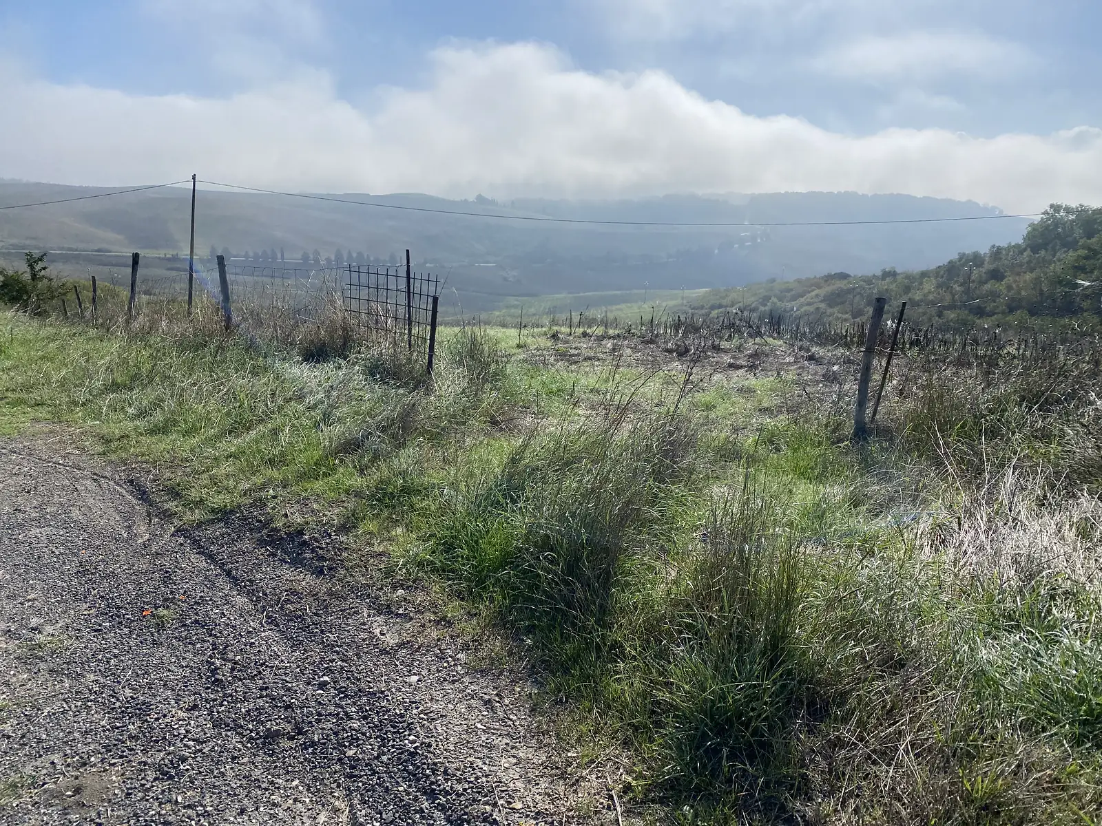 A gravel track running beside a wire fence, overlooking rolling green hills partly veiled in low cloud.