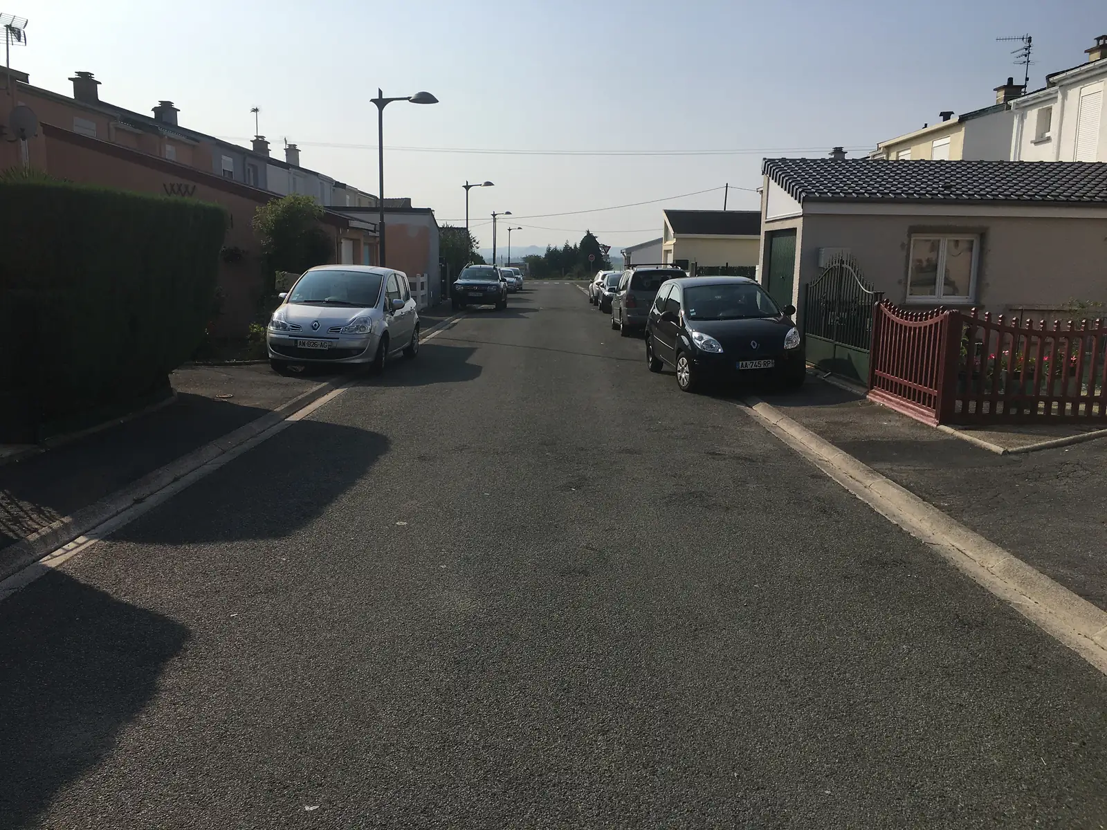 A quiet residential street in a French village lined with terraced brick houses and parked hatchbacks bearing French number plates.