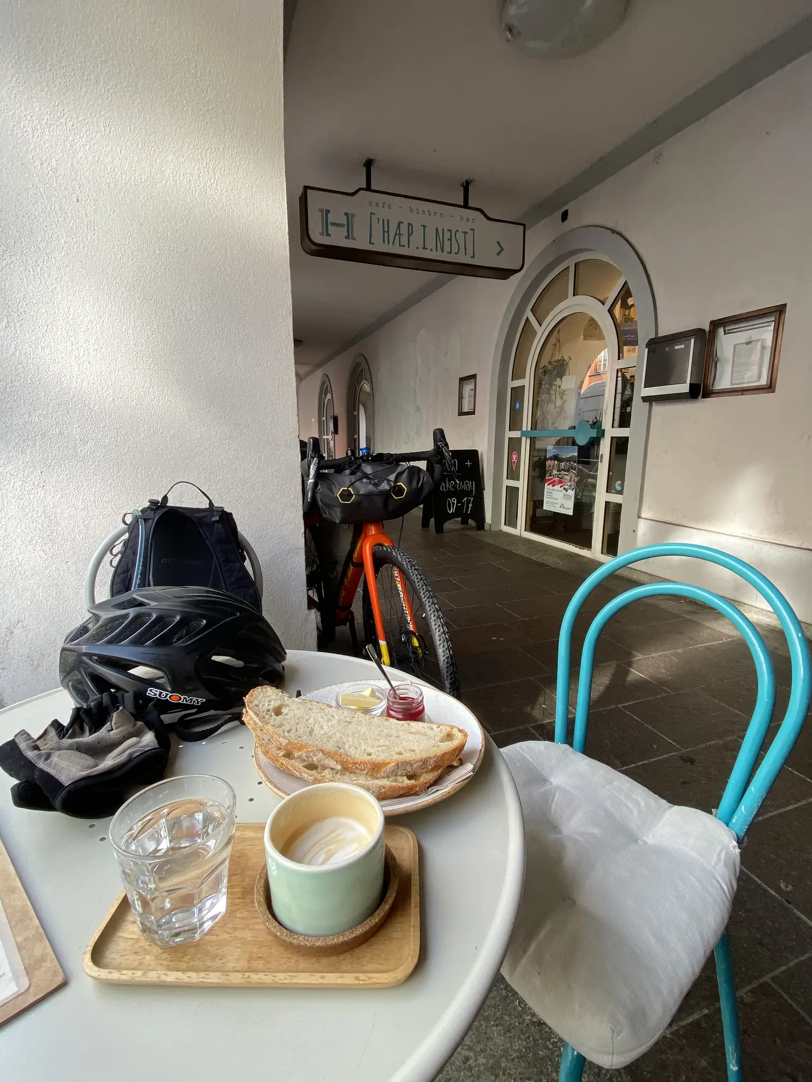 A café table holds a coffee, sliced bread and a helmet beside a loaded touring bike parked outside a café signed 'café - bistro - bar'.