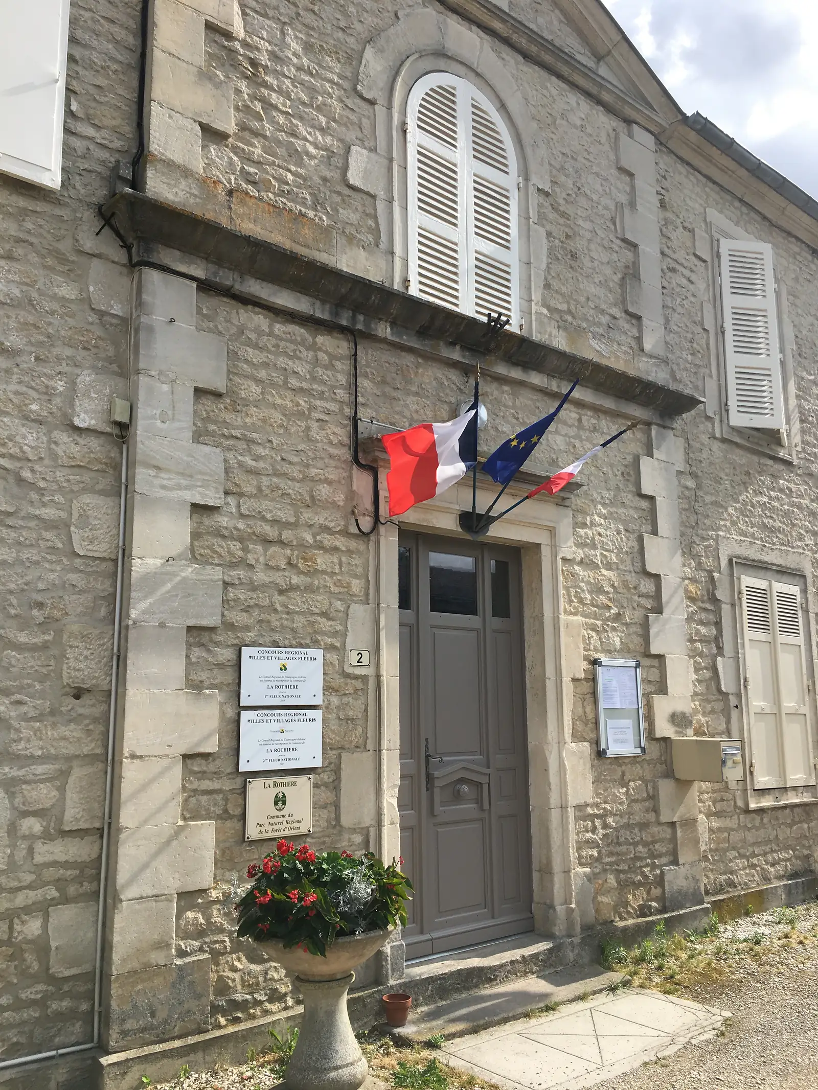 The entrance of La Rothière's mairie, flying French and European flags beside plaques for the Villages Fleuris award and the Parc Naturel Régional de la Forêt d'Orient.