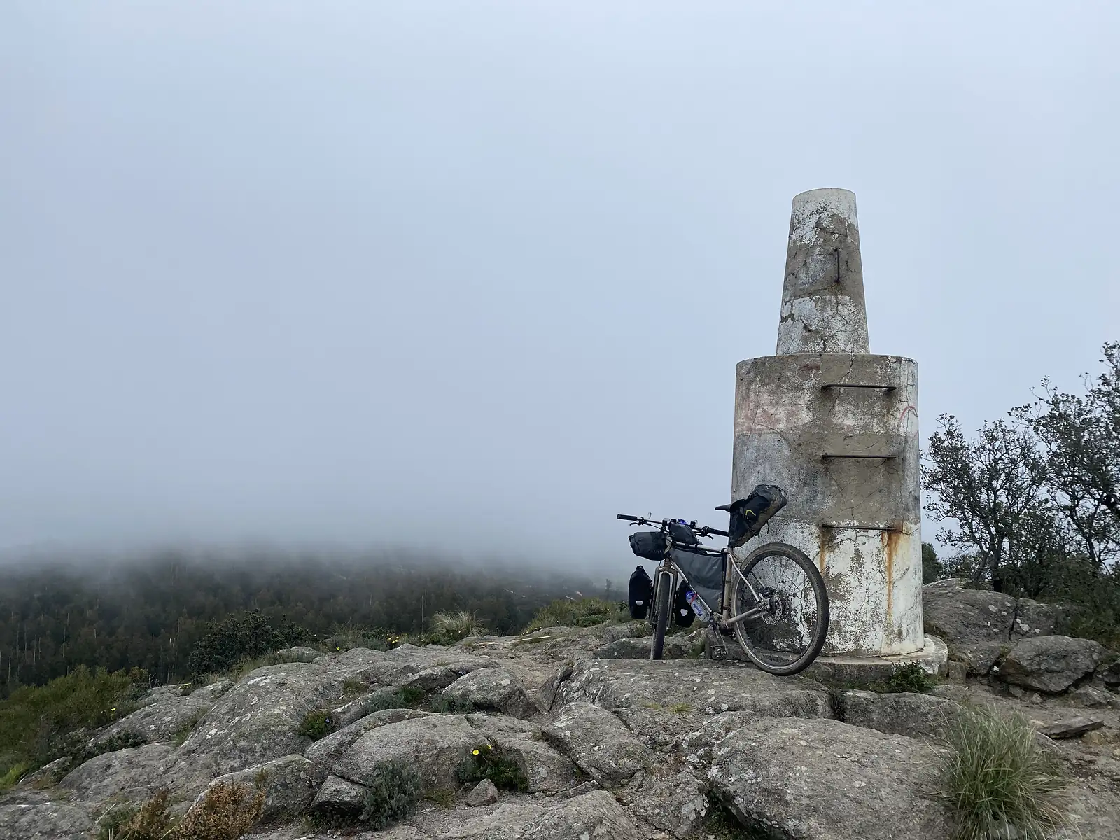 Loaded bike leaning against a squat, weathered concrete summit beacon on bare rock, with cloud banked across the wooded slopes behind.