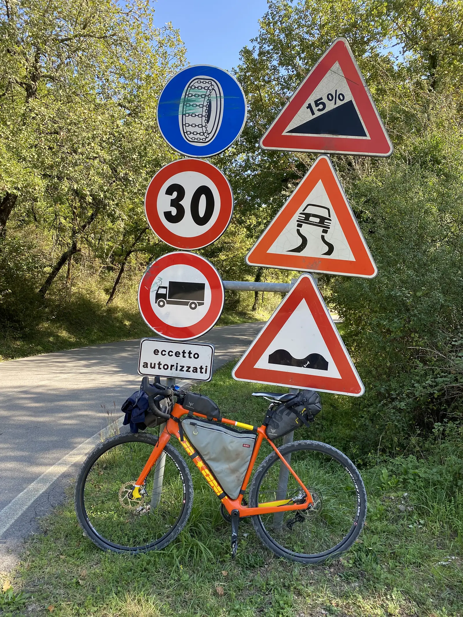 A cluster of Italian road signs warning of a 15% gradient, a slippery bend, a rough surface, snow chains, a 30 km/h limit and a lorry ban marked "eccetto autorizzati", with a loaded orange bike parked beneath them.