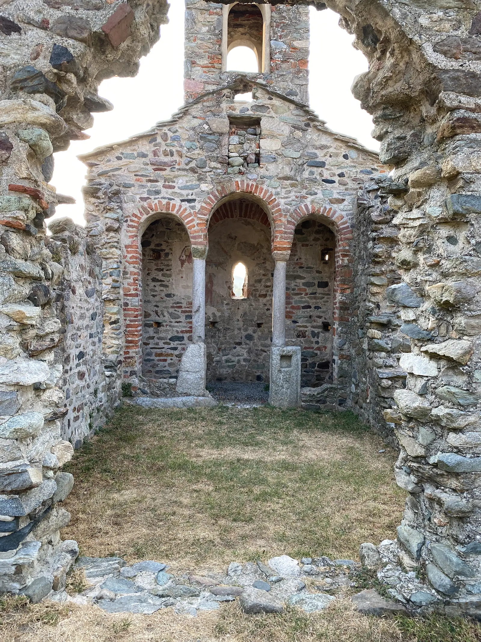 The roofless stone and brick ruins of a small chapel, its arched niches open to the sky above a grassy floor.