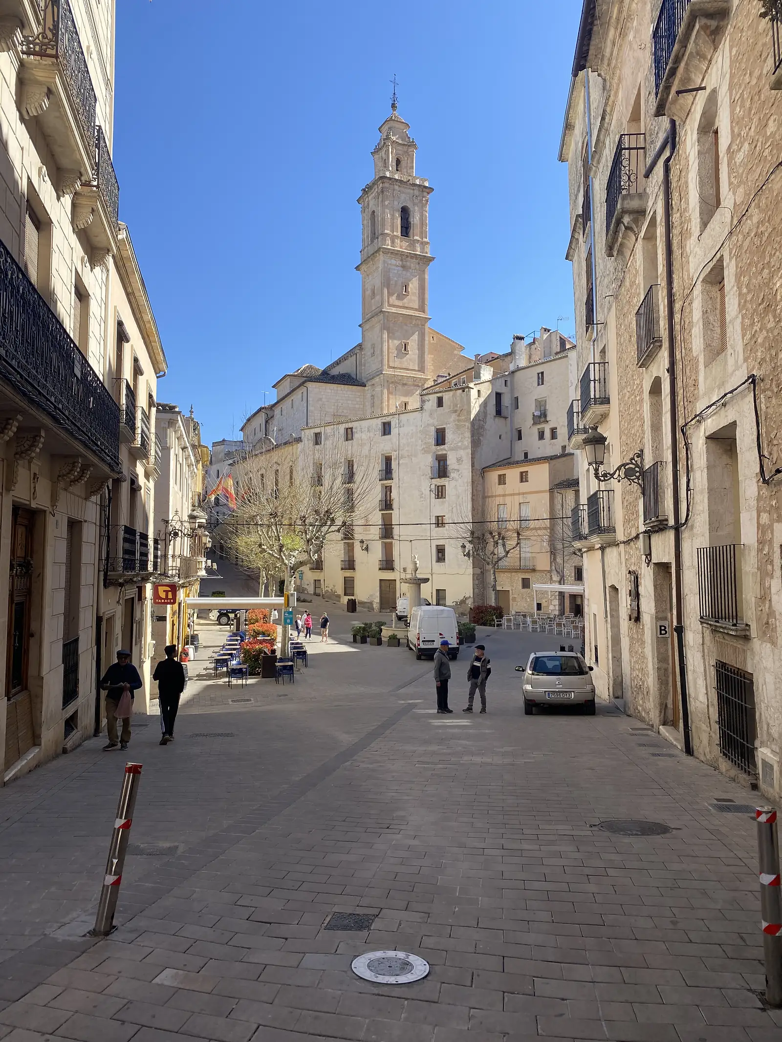 A paved street of stone houses with iron balconies looking down to a small square with cafe tables and a tall baroque bell tower.