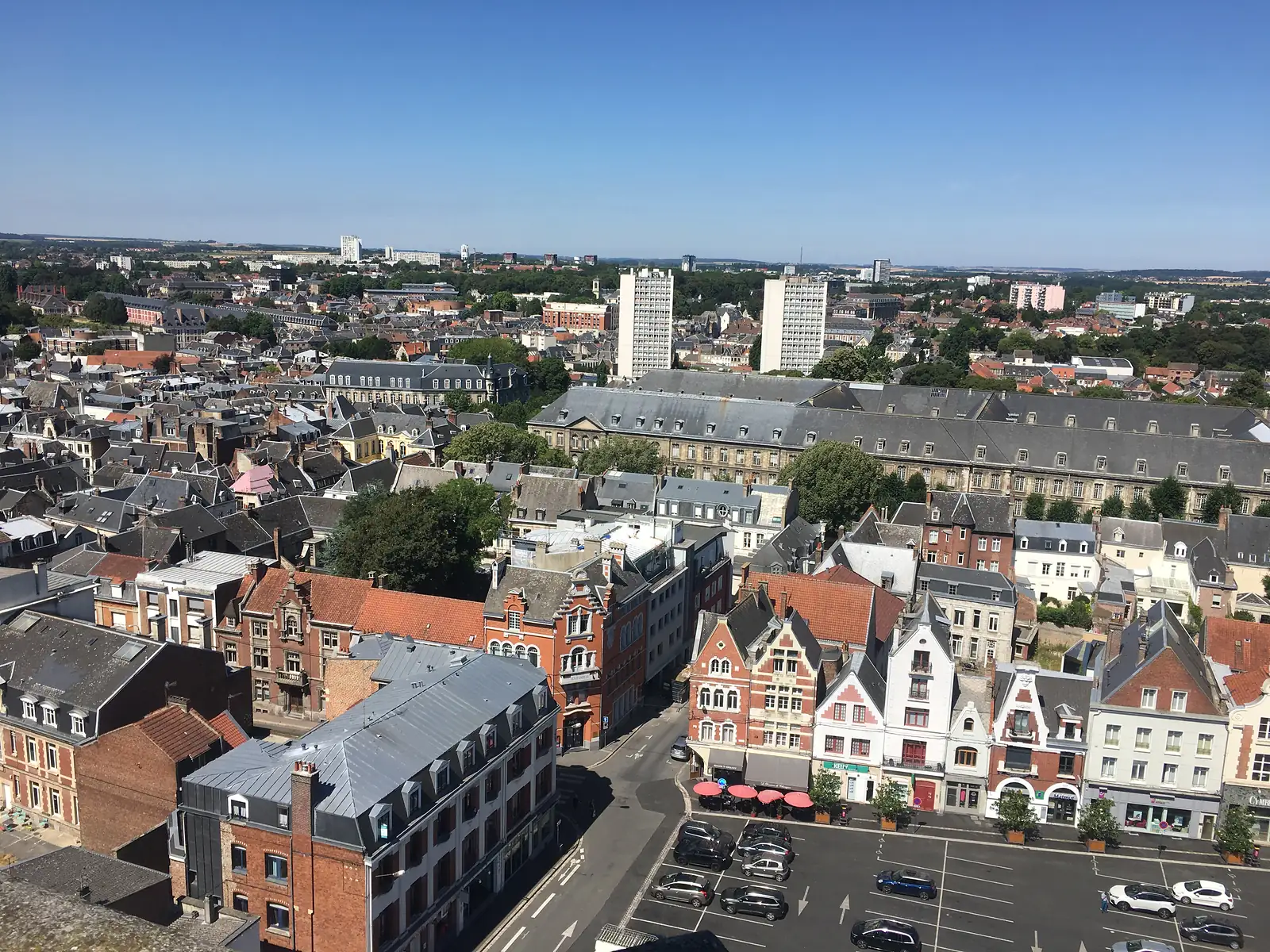 Aerial view over a French town's rooftops from a bell tower, with gabled brick and stone buildings, a red-umbrella café terrace and a car park in the foreground, and two tall tower blocks rising above the wider cityscape under a clear blue sky.