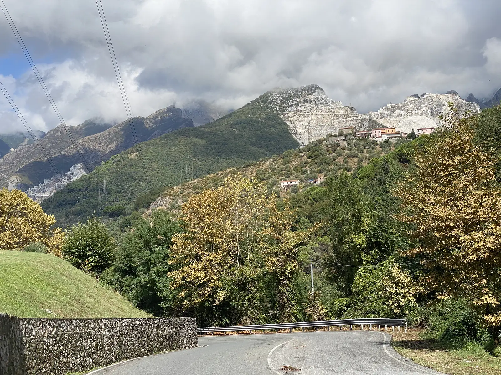 A road curves downhill past a low stone wall and guardrail, with terraced marble quarries streaking a distant mountainside above a hillside village and autumn-tinted trees.