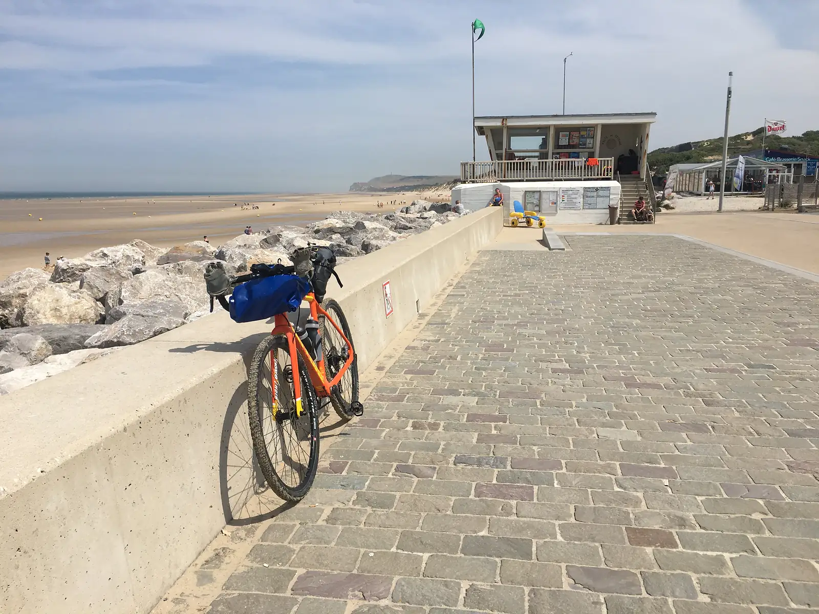 A laden orange gravel bike resting against a stone sea wall near a lifeguard station flying a green flag, with chalk cliffs visible along the beach beyond.