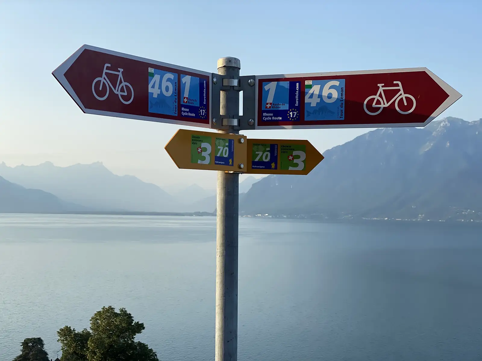 A signpost above the lake shows red cycle-route arms for "46 Tour du Léman" and "1 Route du Rhône / EuroVelo 17" crossed with yellow ViaFrancigena arms marked "3 Chemin panorama alpin" and "70".