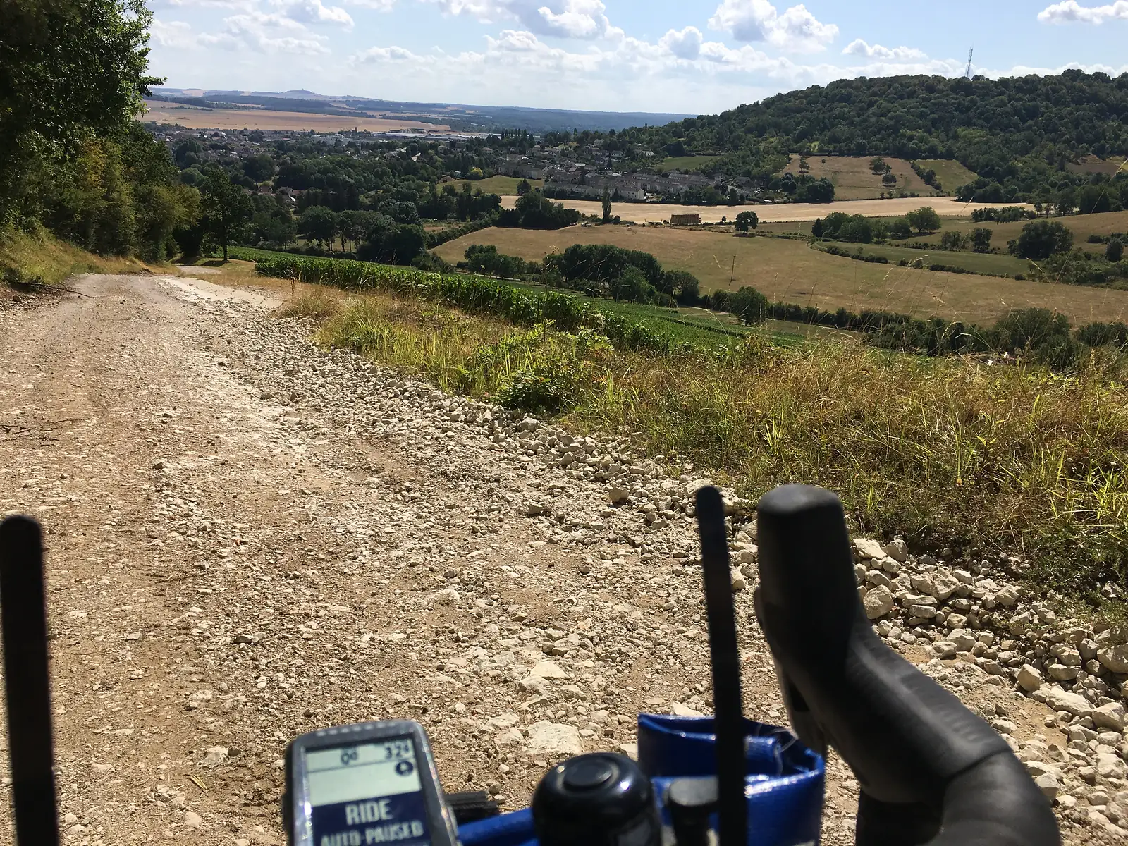 A chalky gravel track drops away downhill towards a town in the valley below, with a cycling computer reading 'RIDE AUTO-PAUSED' visible on the handlebars.
