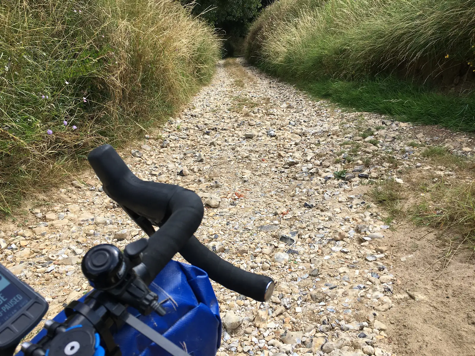 A flint-strewn gravel track stretches ahead between tall roadside grasses, seen over a bicycle's drop handlebars and blue bikepacking bag.
