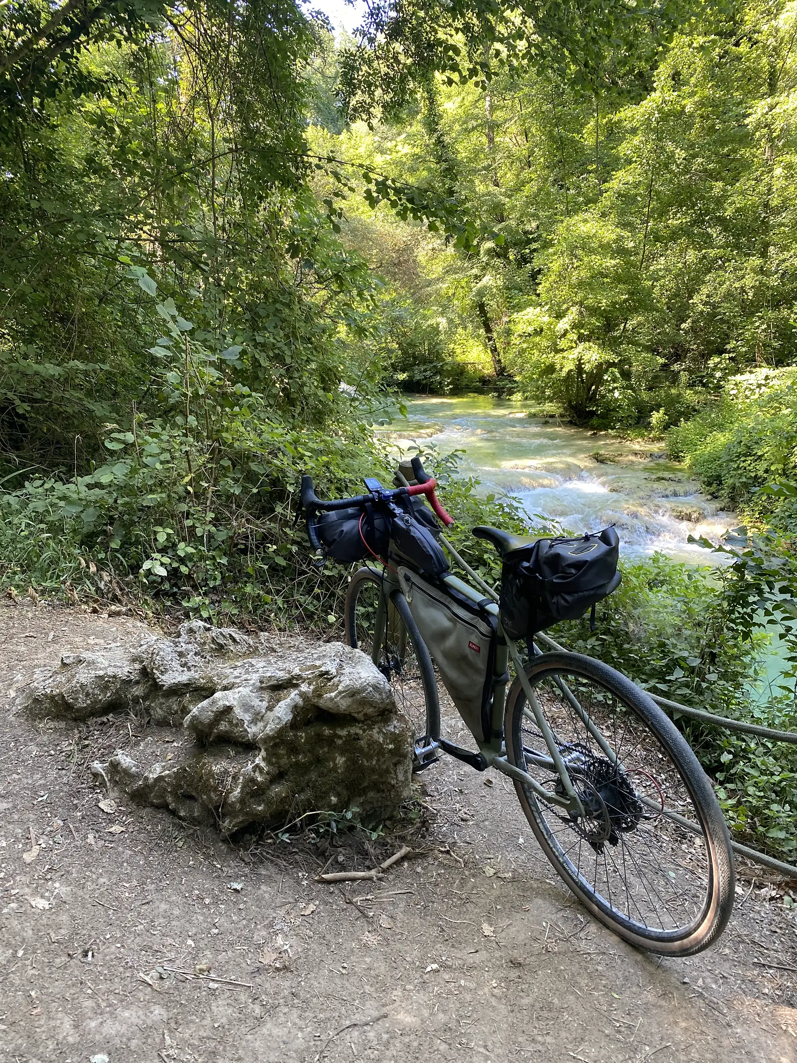 A drop-bar touring bike loaded with bikepacking bags leans against a rock beside a wooded stream running over small cascades.