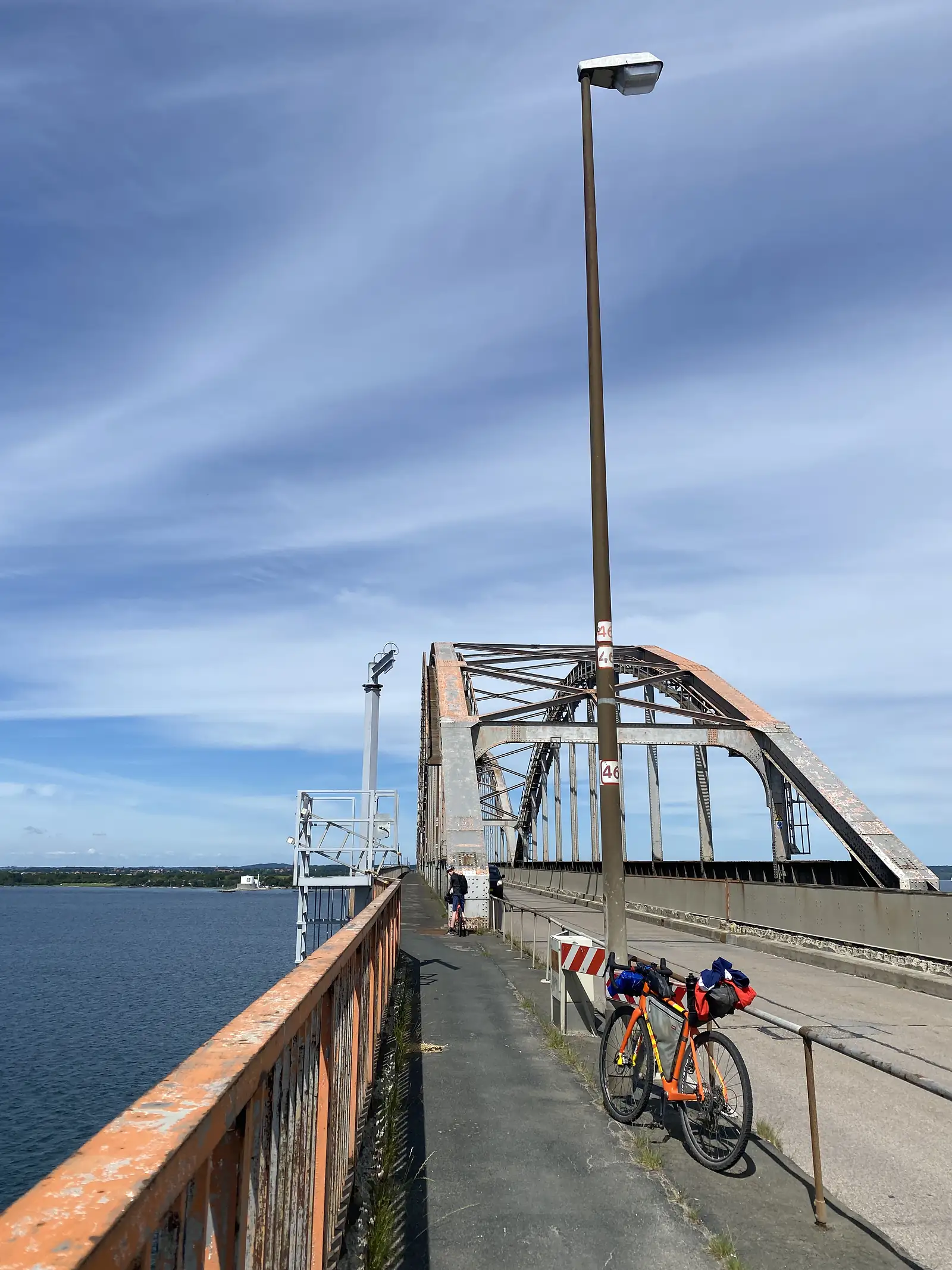 A loaded orange bike propped by a lamp post on a causeway bridge's cycle path, with a rusting steel arch span ahead and a rider standing beyond.