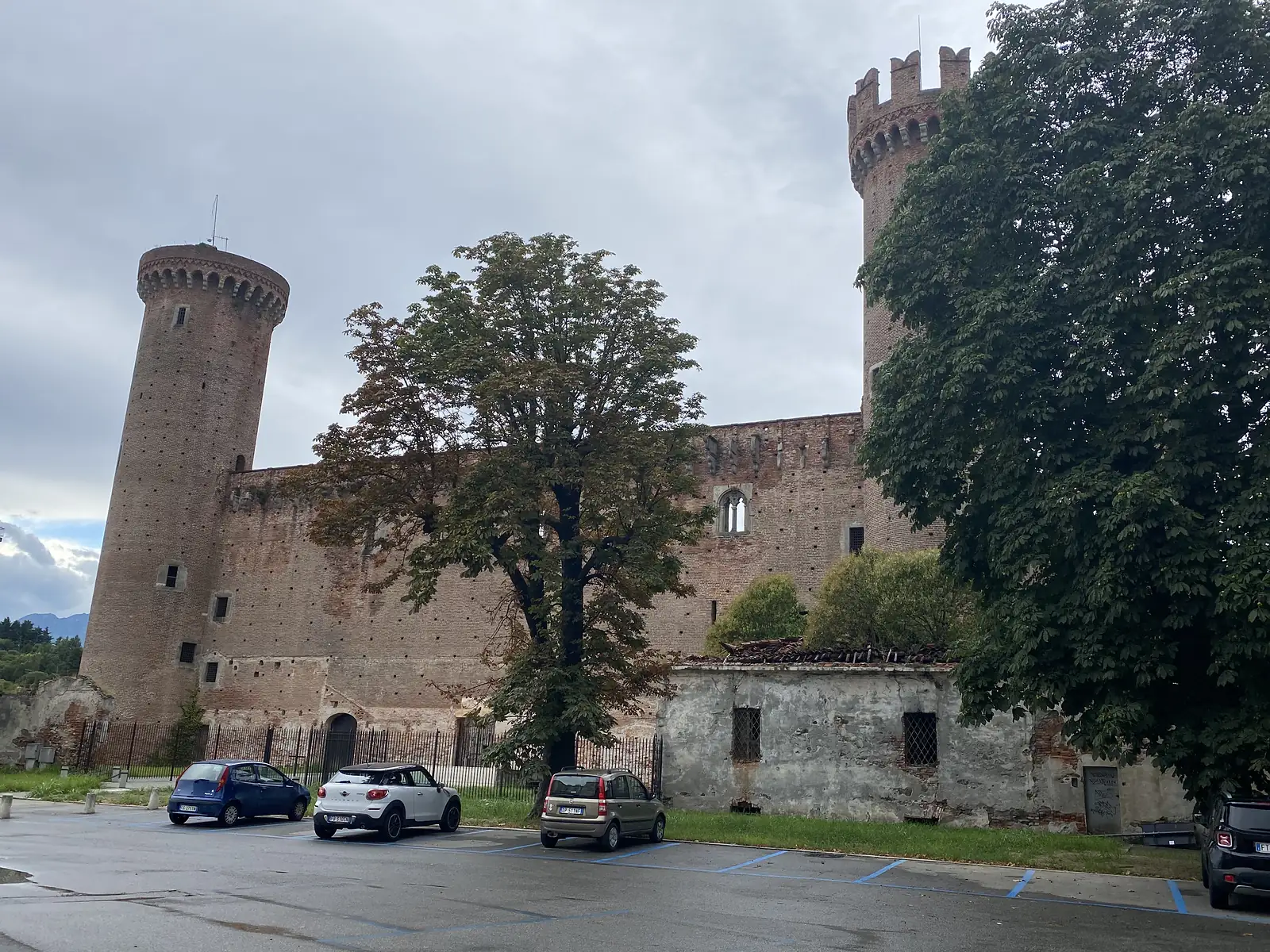 The brick towers and battlemented curtain wall of Ivrea Castle rise above a car park, one round tower topped with a crenellated crown and a tall square tower behind.