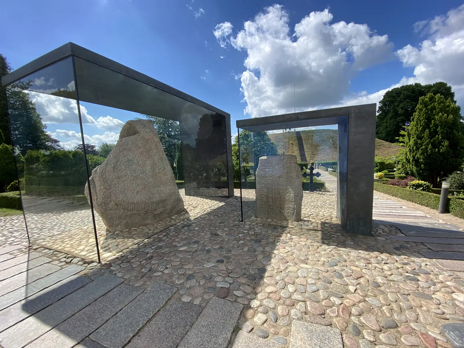 Two carved rune stones sheltered under angular glass and steel canopies on a cobbled churchyard path, with a green burial mound behind.