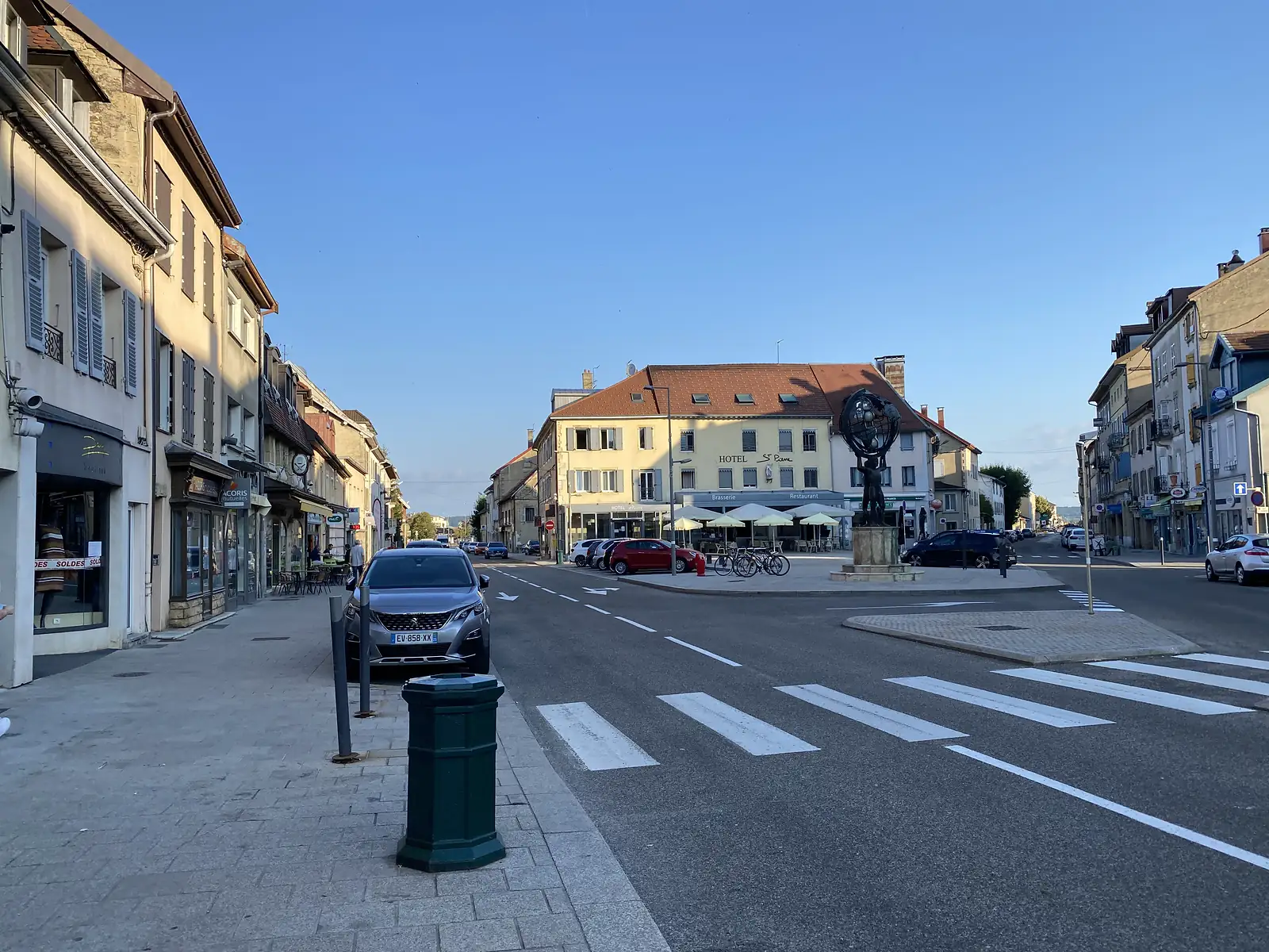 A market square in a French town, with a bronze Atlas statue supporting a globe outside the yellow-fronted Hotel St Pierre, café tables, parked cars and a zebra crossing in the foreground.